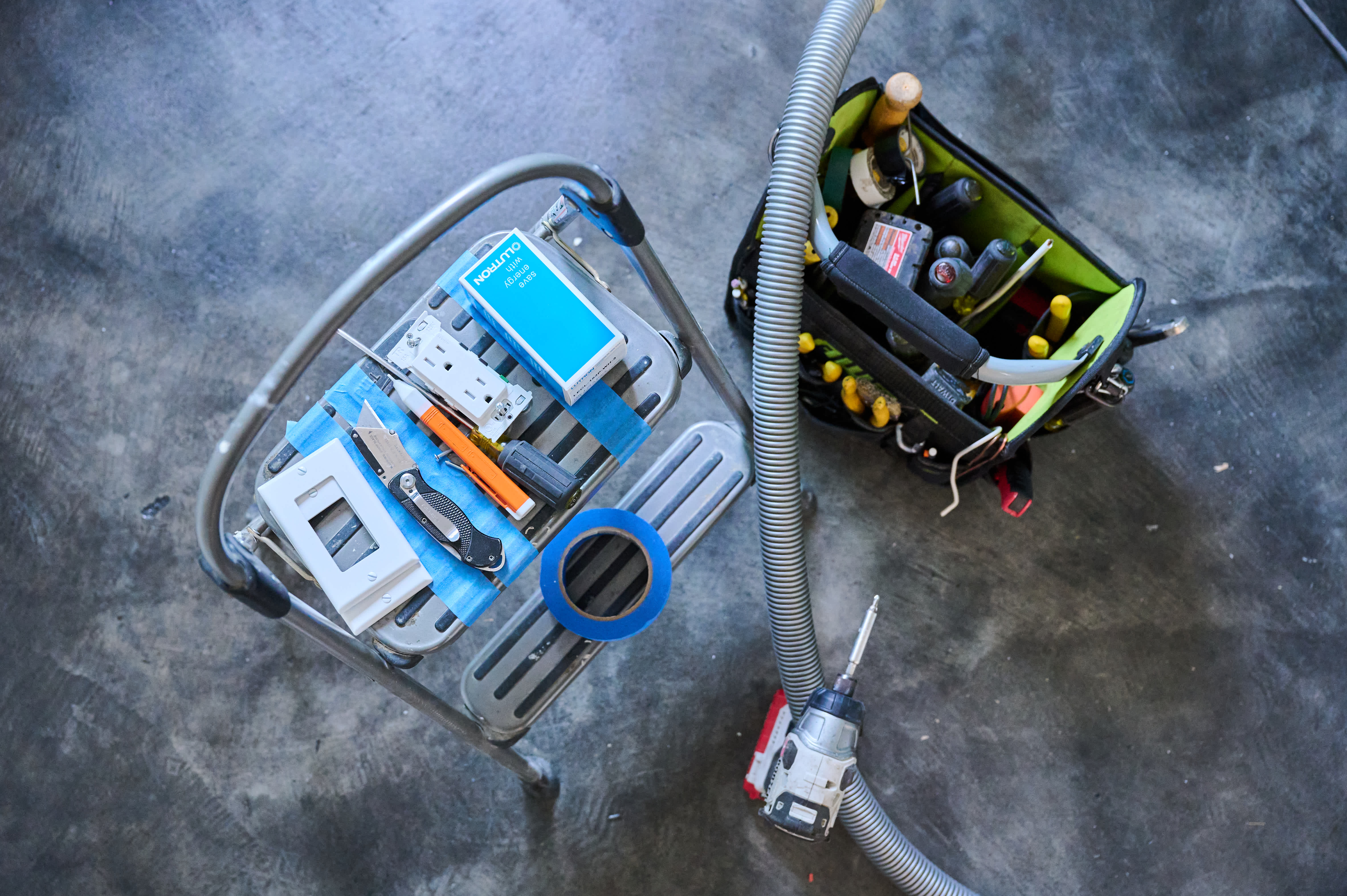 A step stool holds electrical tools, outlet covers, and tape, while a tool bag and drill sit nearby, indicating electrical work in progress.
