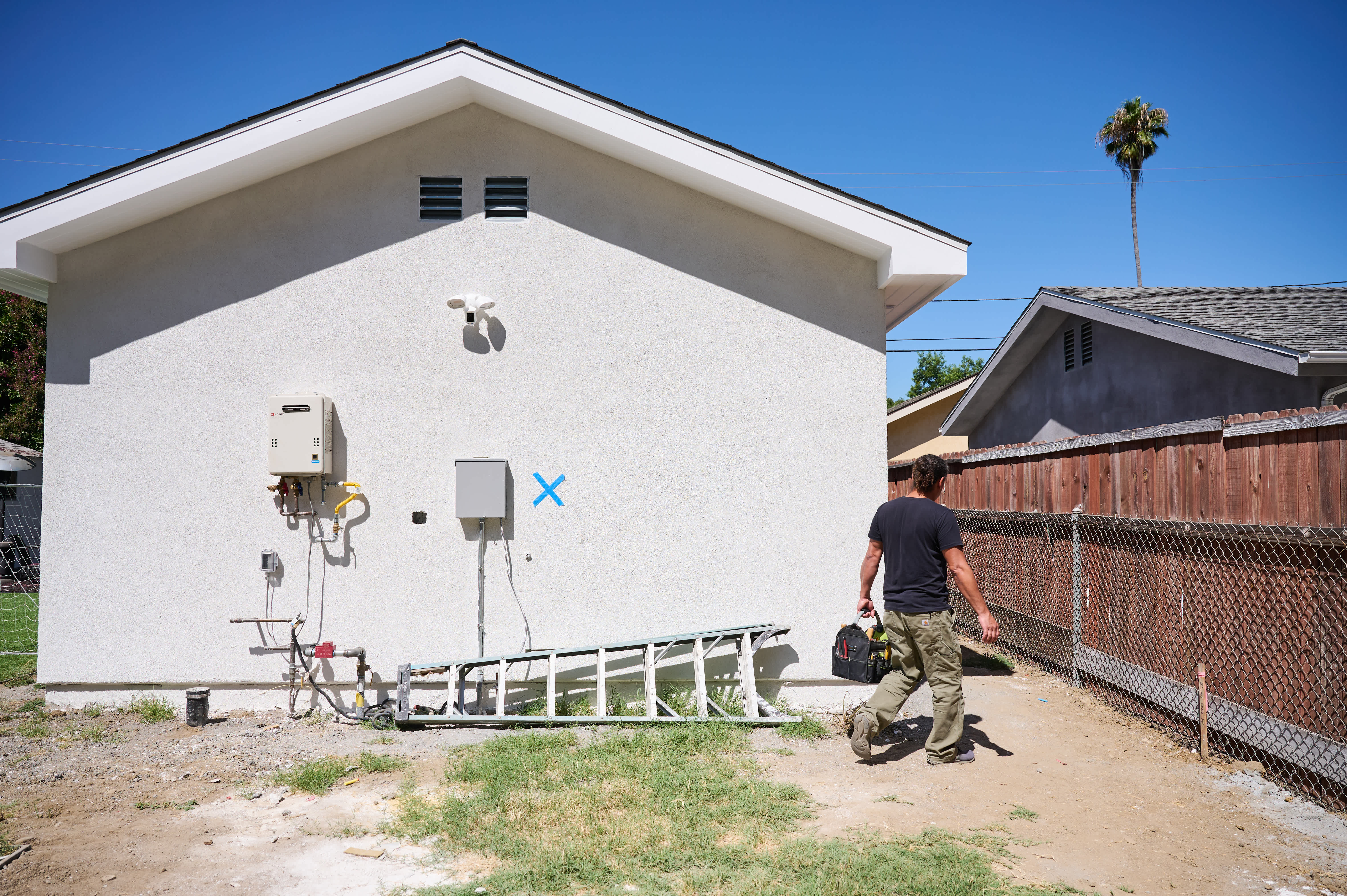 A technician in a black shirt and khaki pants walks away from a house wall with utility boxes, a ladder, and a blue \"X\" marking on it.