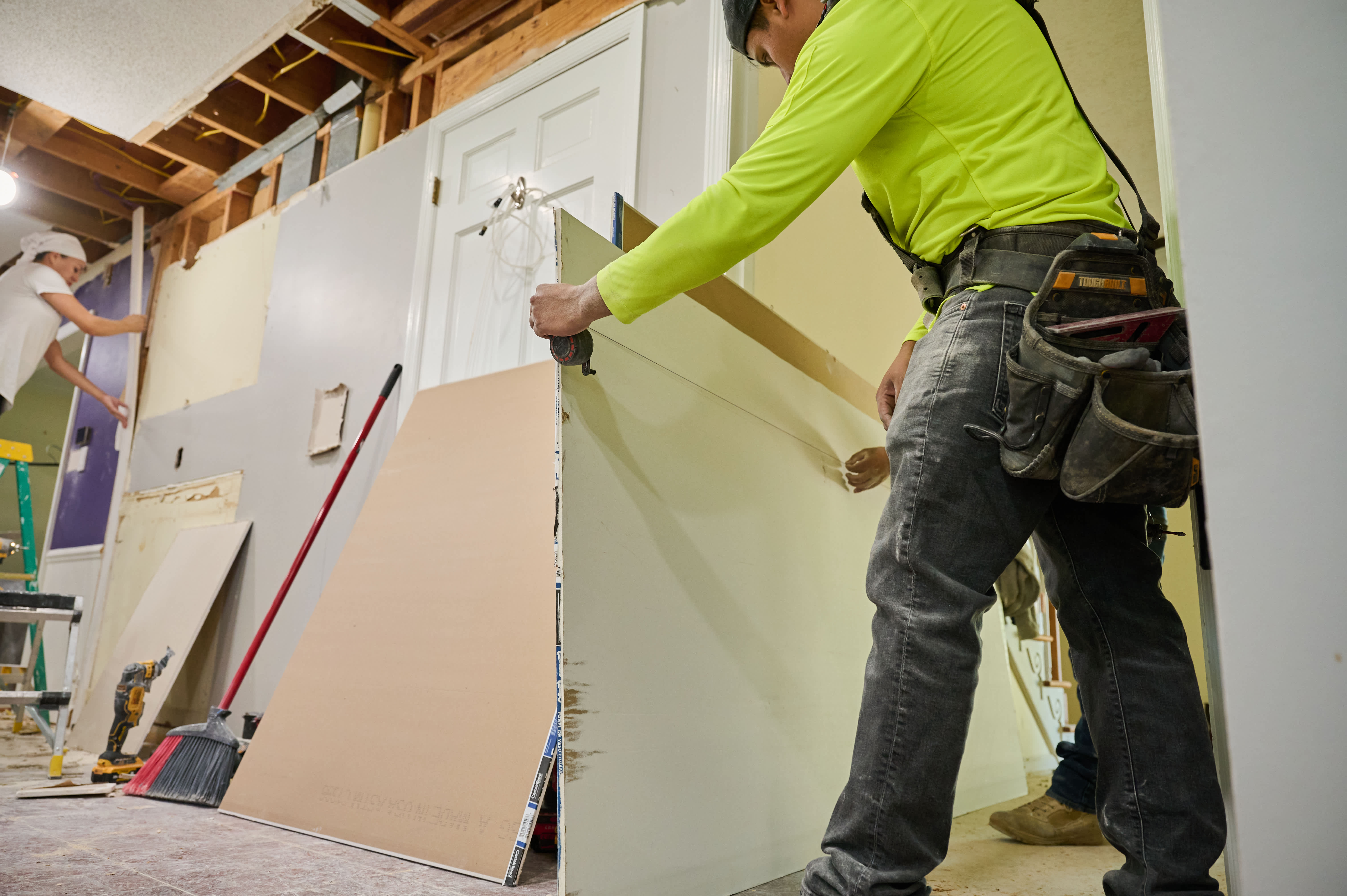 A construction worker measuring drywall while another adjusts its position during a home renovation project.