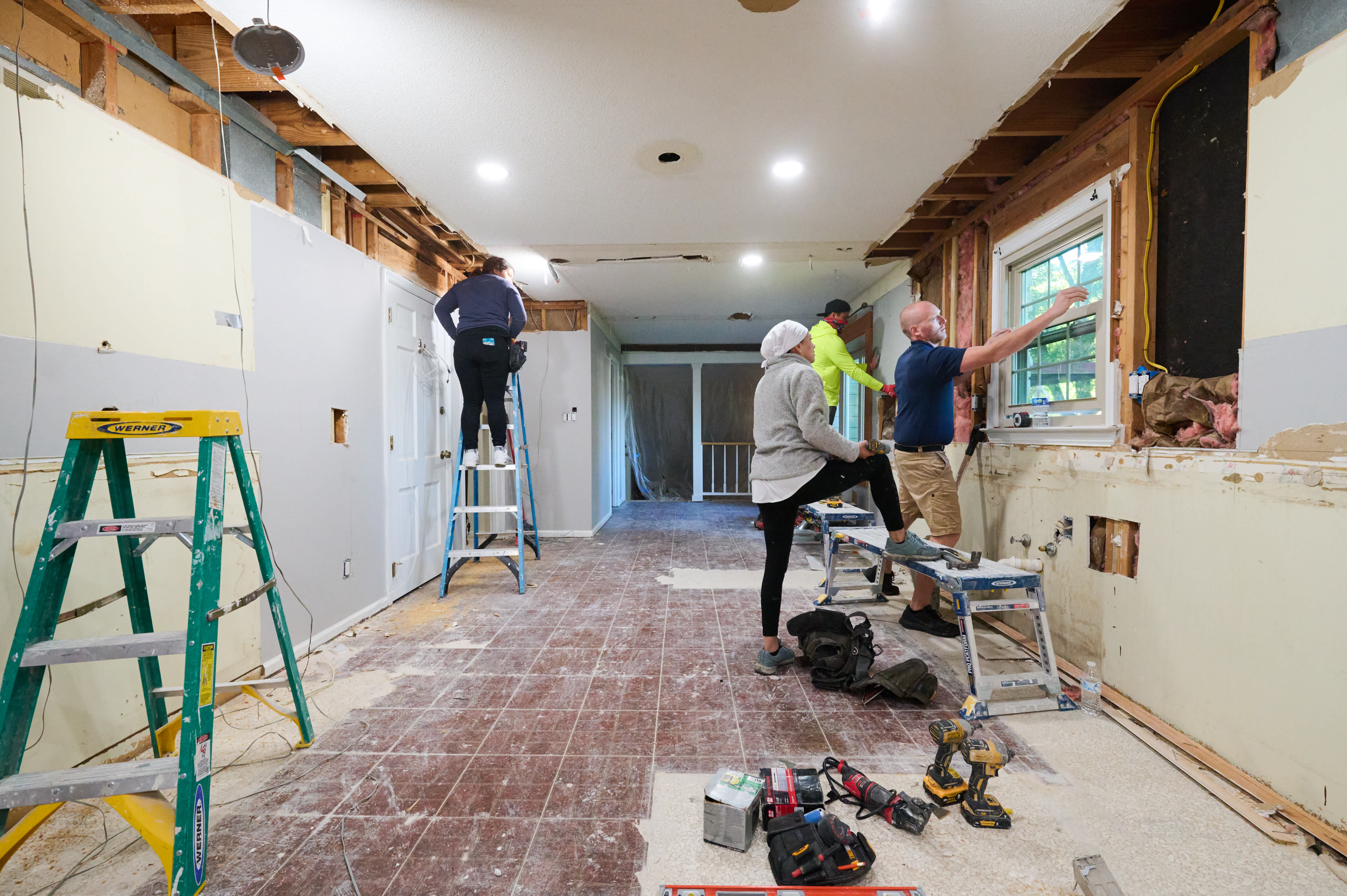 A renovation team works on walls and ceiling; one person is on a ladder while others remove drywall near a window and inspect progress.