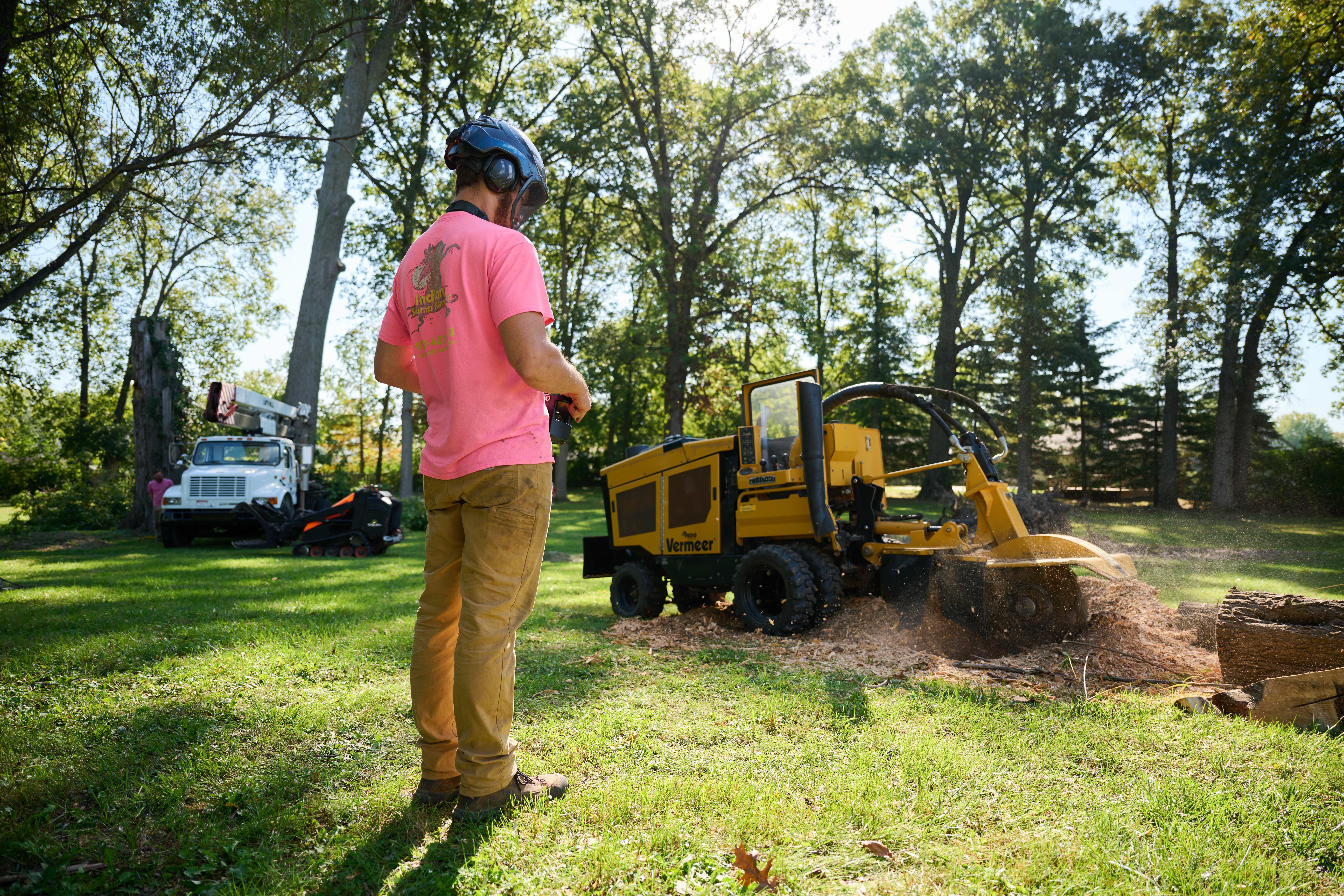 Tree worker in safety helmet operates a stump grinder to remove a tree stump in a grassy yard with trucks in the background.