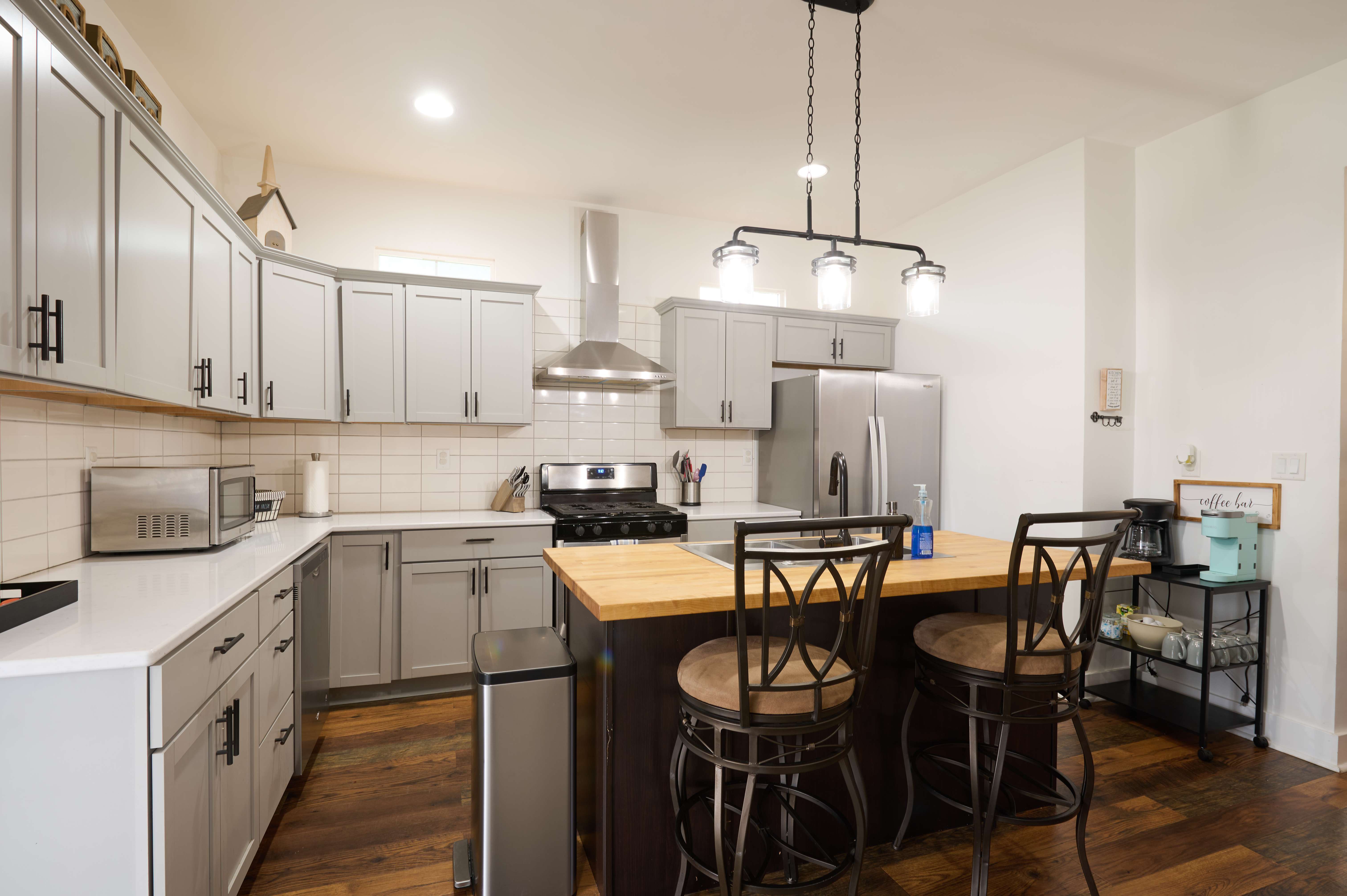A modern kitchen with gray cabinets, a center island with wooden countertop and bar stools, stainless steel appliances, and pendant lighting.