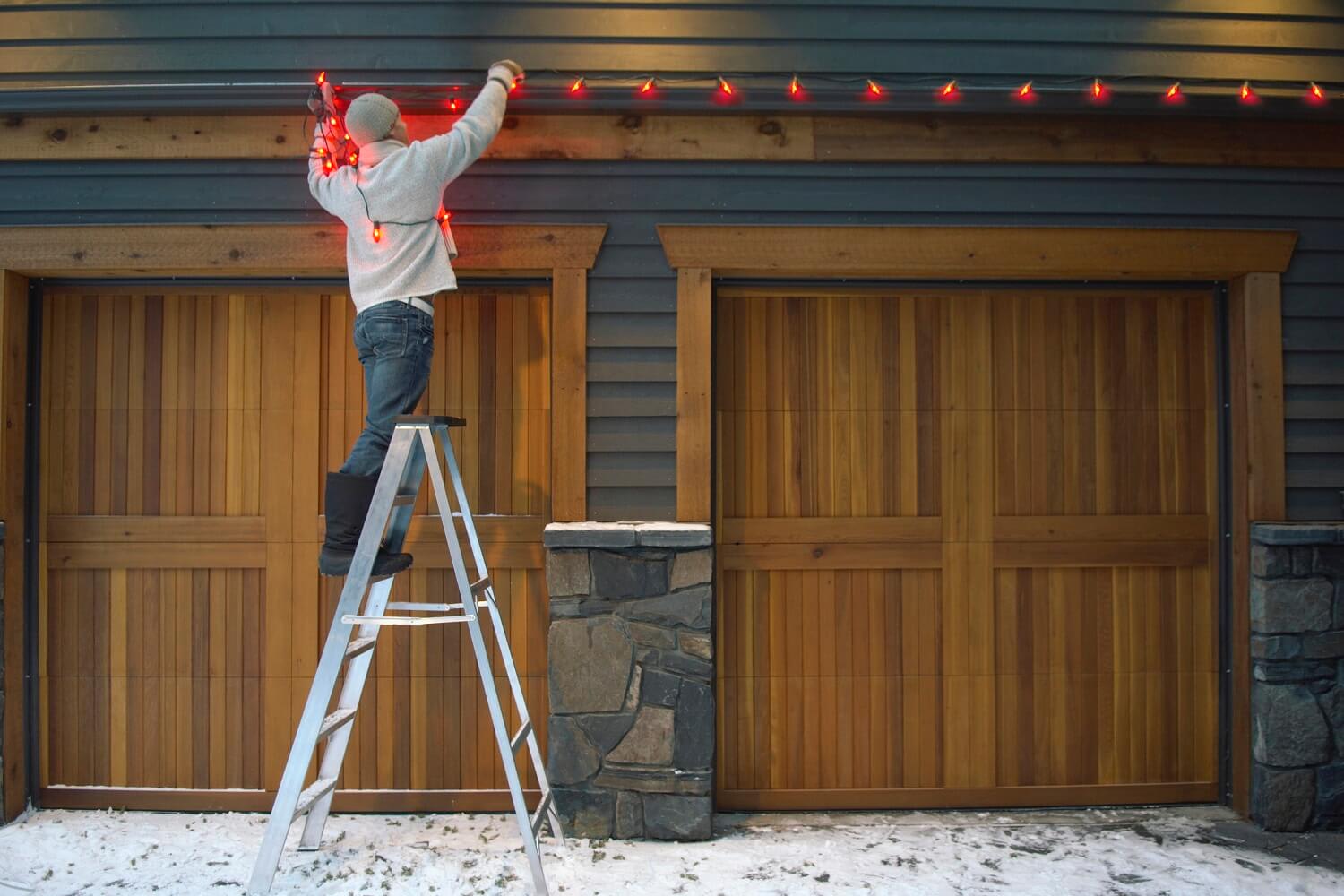 Man standing on ladder hanging holiday lights on top of the garage