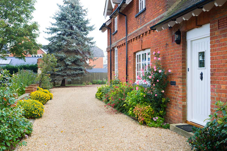 gravel driveway lined with lots of plants and shrubbery and a red brick house and white door on the other side