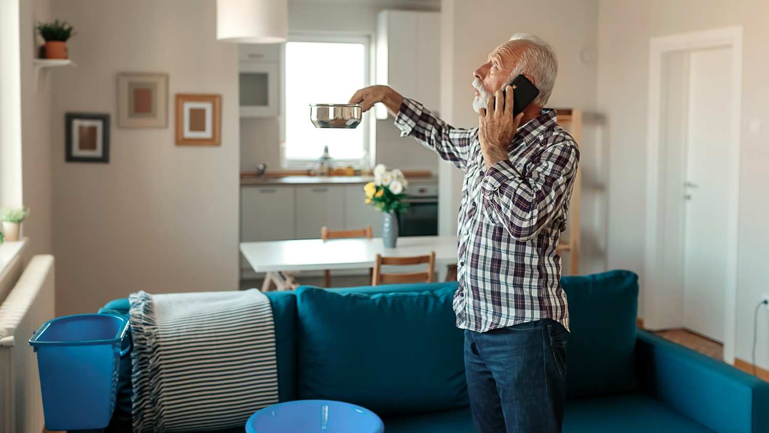 text:Angry Bearded Senior Man Talking on Smartphone Near Plastic Wash Bowl at Home in the Living Room Because of Roof Leaking