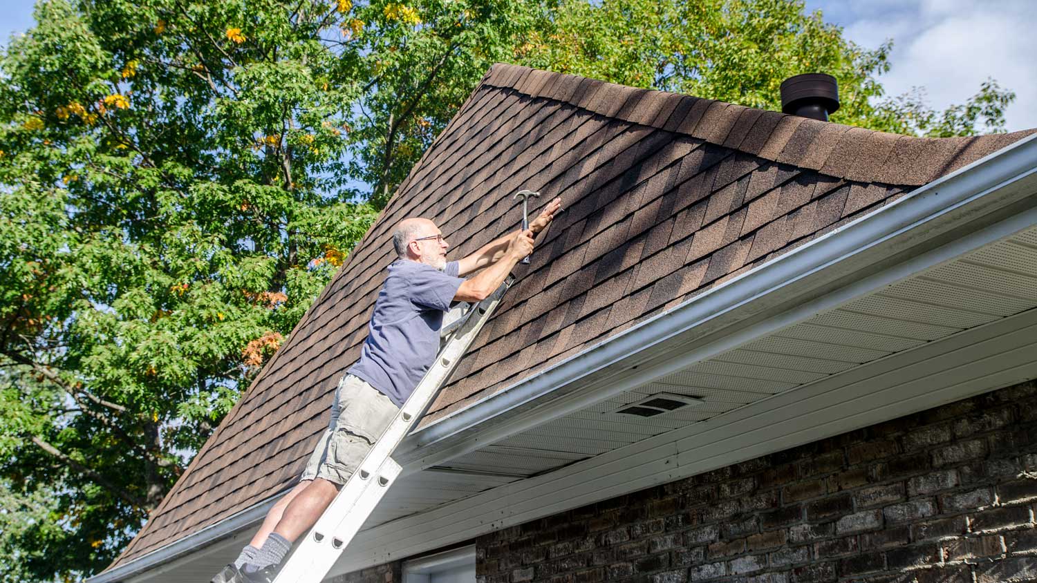 Mature man standing in ladder and holding asphalt shingle to repair roof during summer day