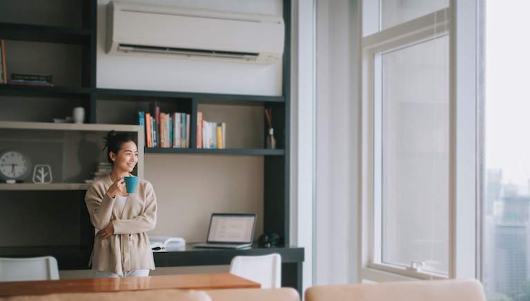 woman with cup of coffee near large windows and underneath air conditioning vent