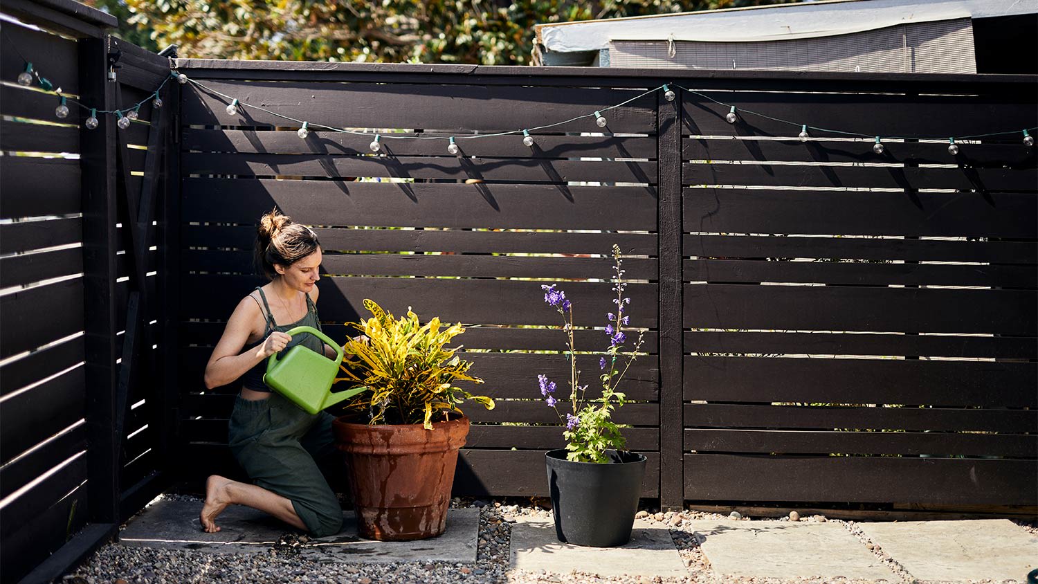 Woman watering potted plant while kneeling by fence at backyard