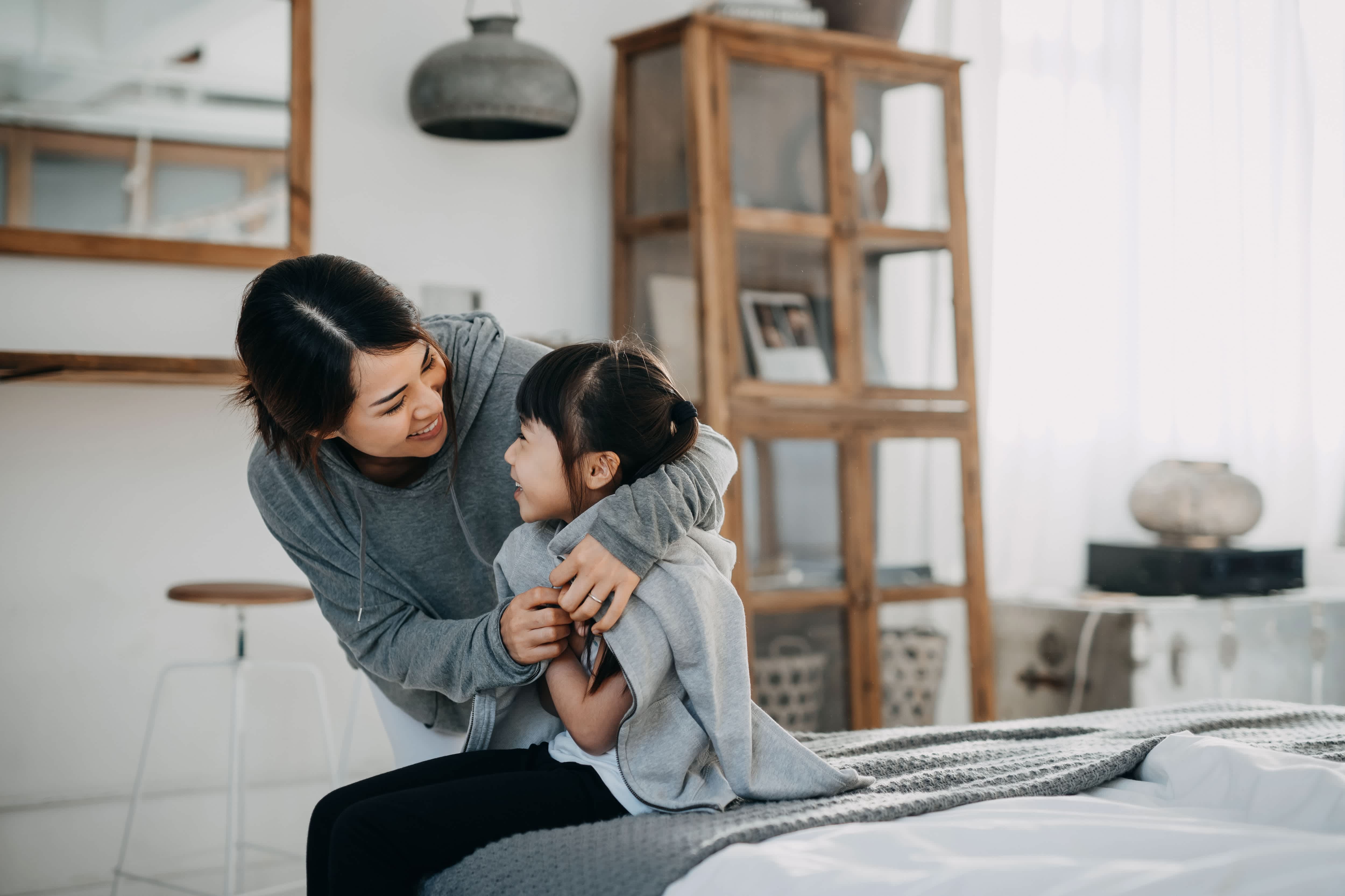 Young mother draping coat over her daughter’s shoulders inside their home