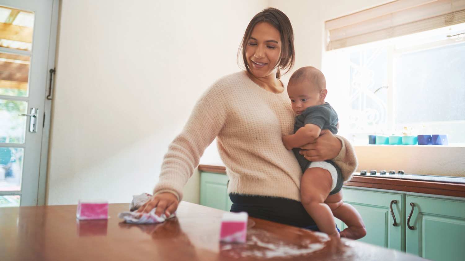 young woman carrying her baby son while cleaning the kitchen at home