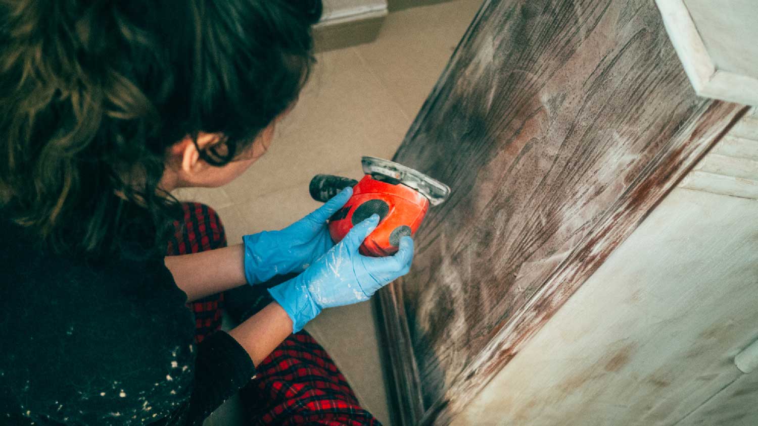 A woman sanding a wooden cabinet with an orbital sander