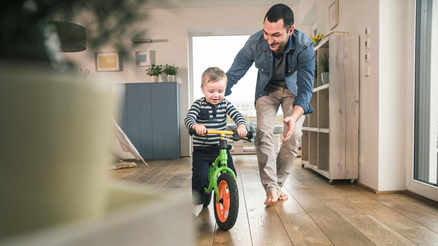 A father helping his son to balance bicycle in a room with hardwood floors
