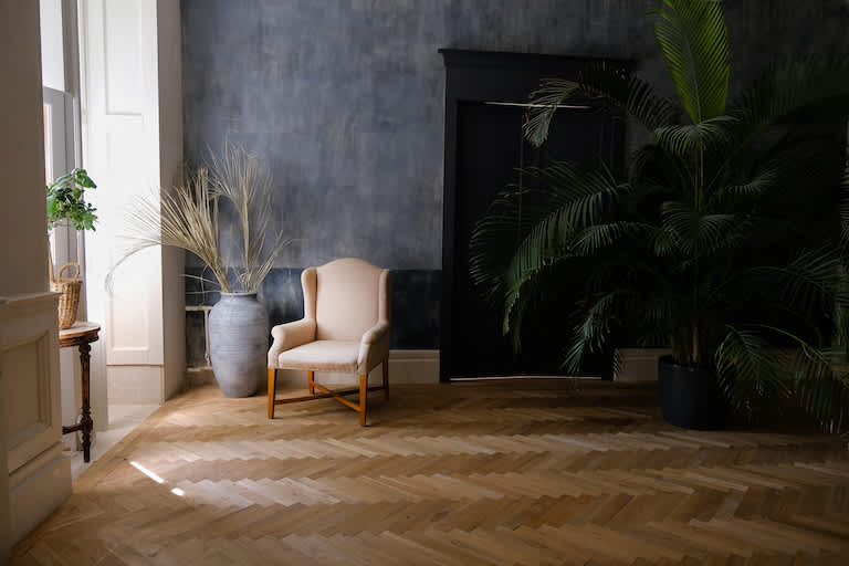 off-white accent chair sits in corner by window in room with dark blue walls and diagonal plank wood floors