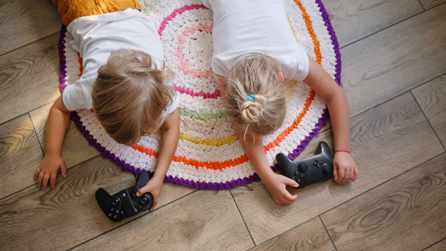 Two siblings laying on a rug on a laminate floor playing videogames