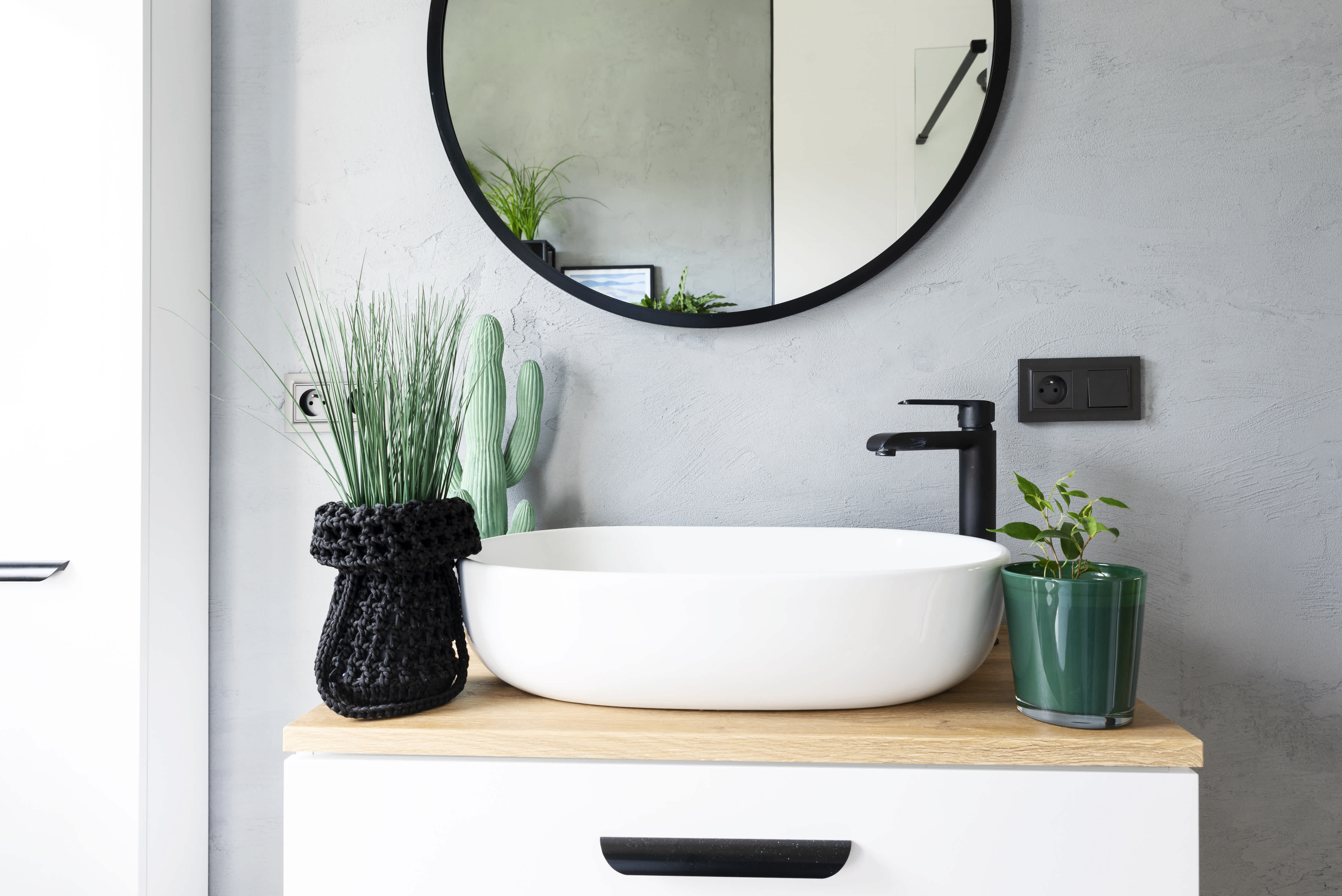 Small vessel sink with black faucet and green plants underneath a round black mirror