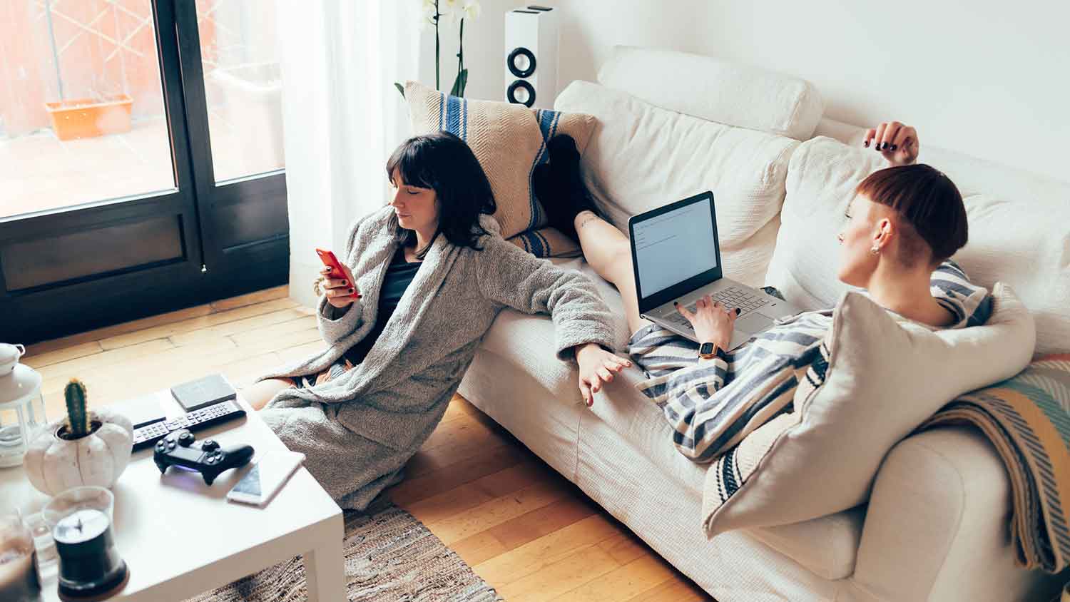 Two women sitting on the couch checking their phone and laptop