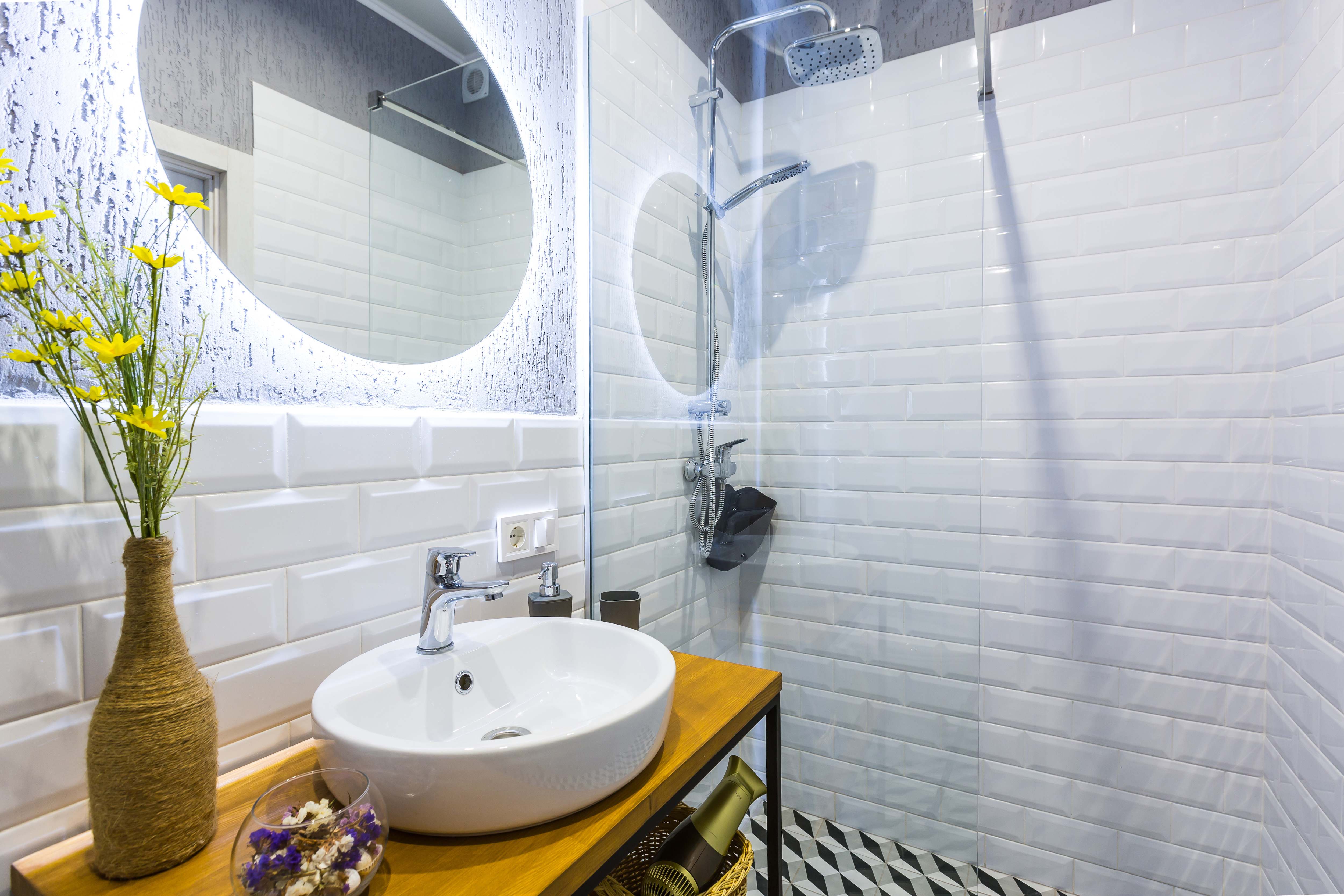 Modern small bathroom with white tile walls, glass rain shower, backlit round mirror, and vessel sink on narrow wood table