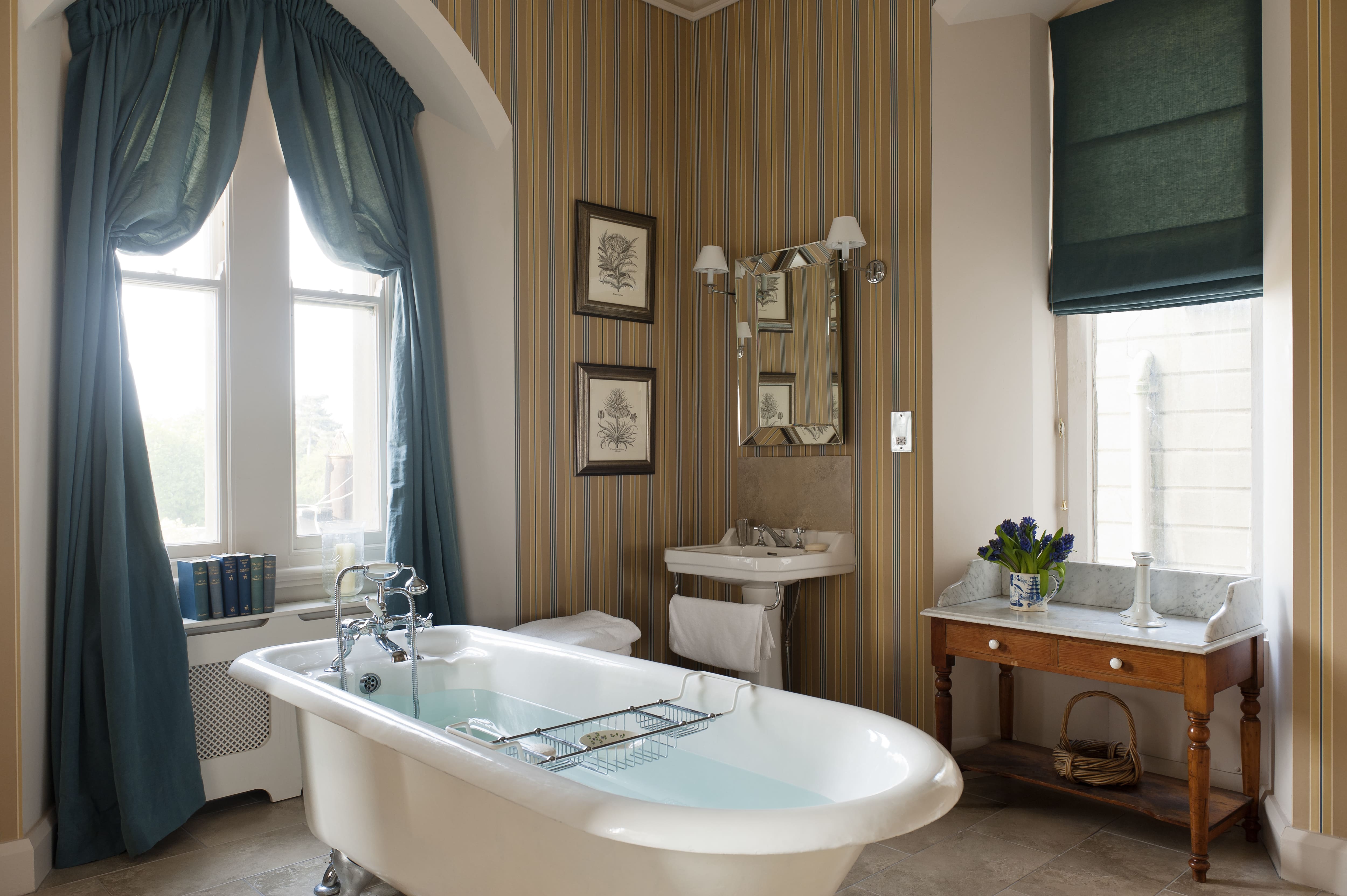 Bathroom with freestanding roll-top tub, teal and brown striped wallpaper, pedestal sink, brown vanity, and teal curtains