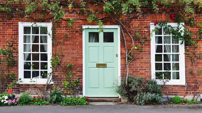 closeup of home exterior with brick walls, green front door, and two windows on either side, as well as plants around doorway