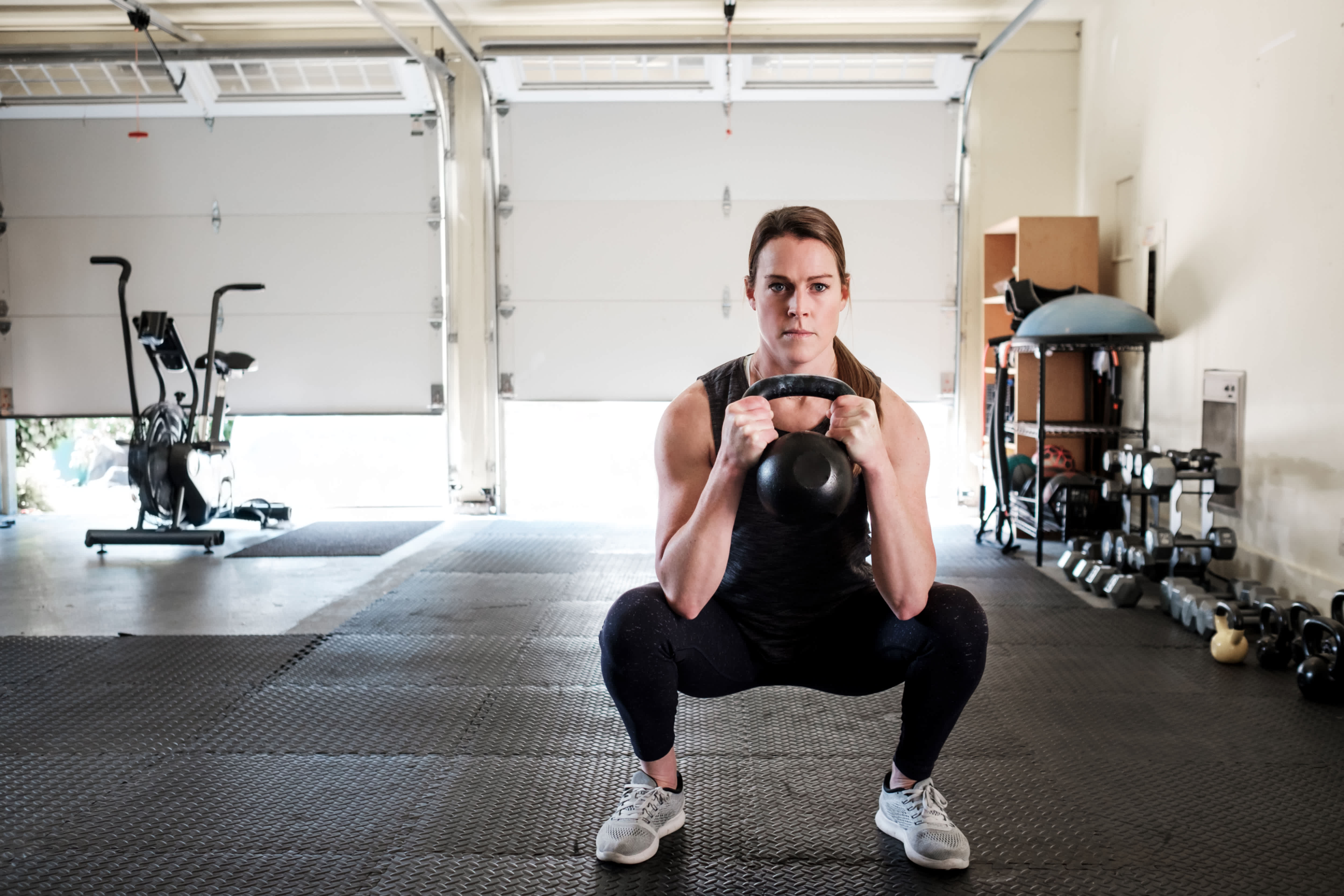 Woman in gym clothes is doing a squat with a kettlebell in her garage home gym with black rubber interlocking tiles on the floor