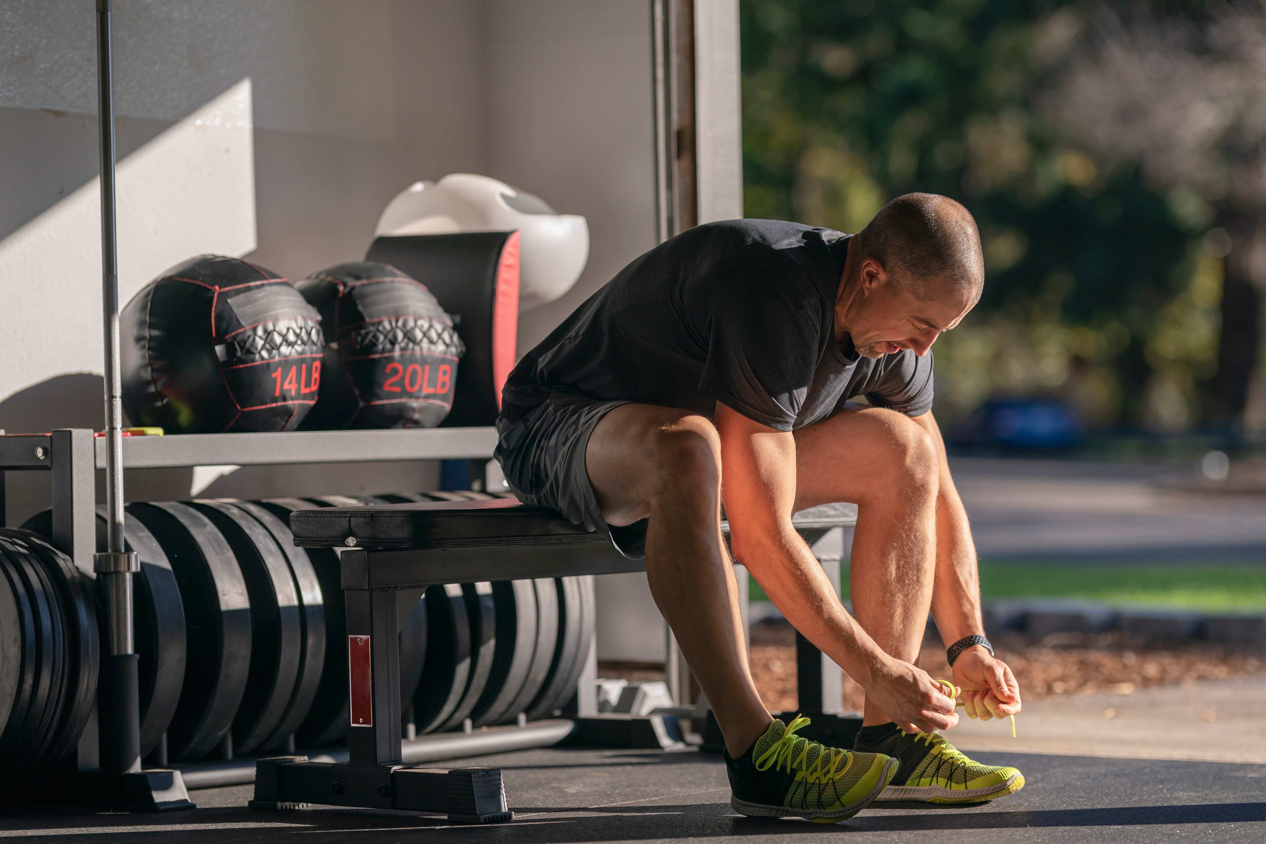 Man in gym clothes lacing up his sneakers in his garage home gym with gym equipment stored behind him