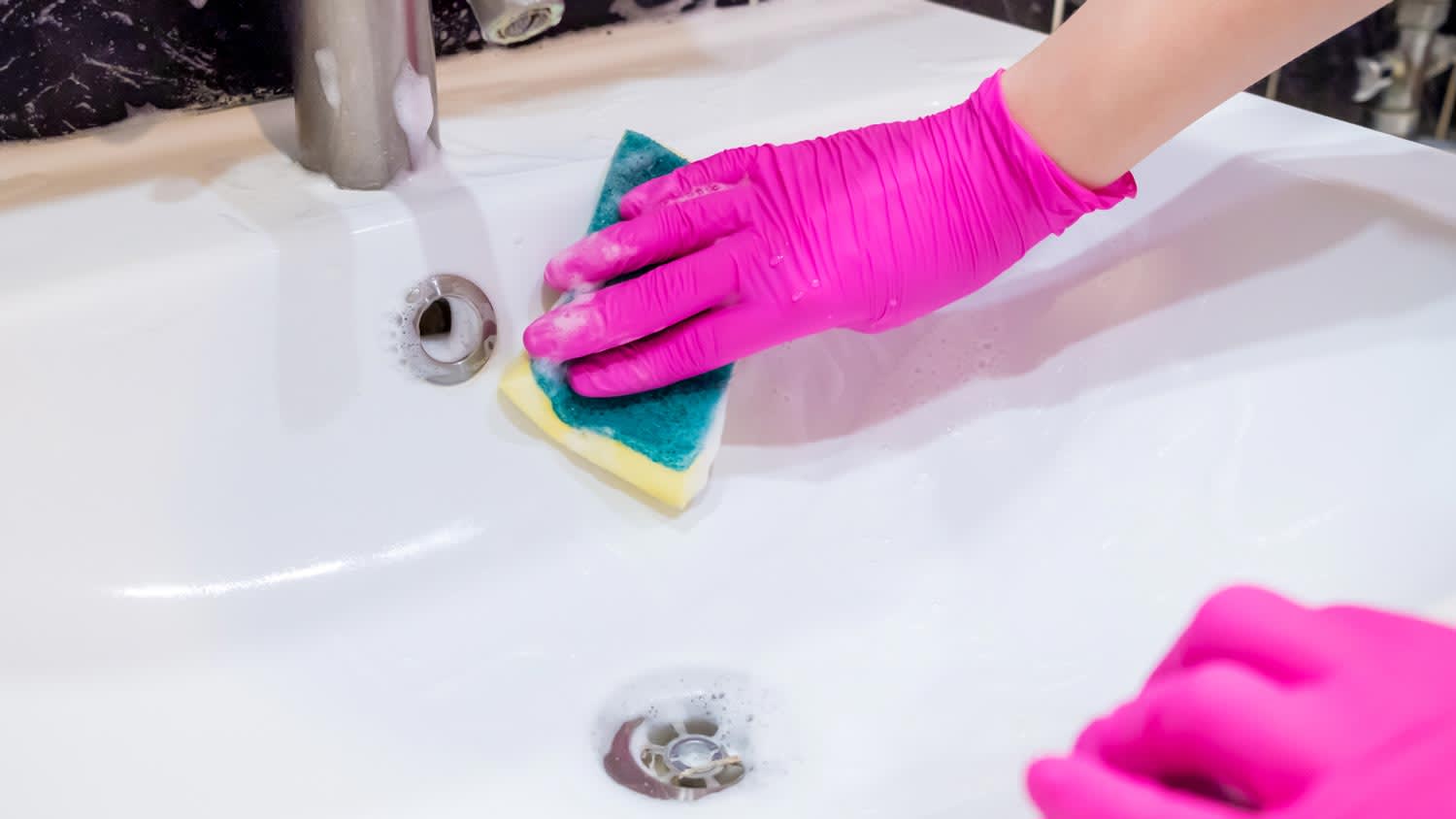 A woman cleaning a bathroom sink using a sponge and bleach