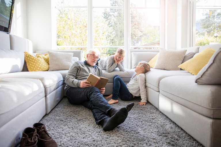 elderly man and two young kids read book in front of large windows between two white couches