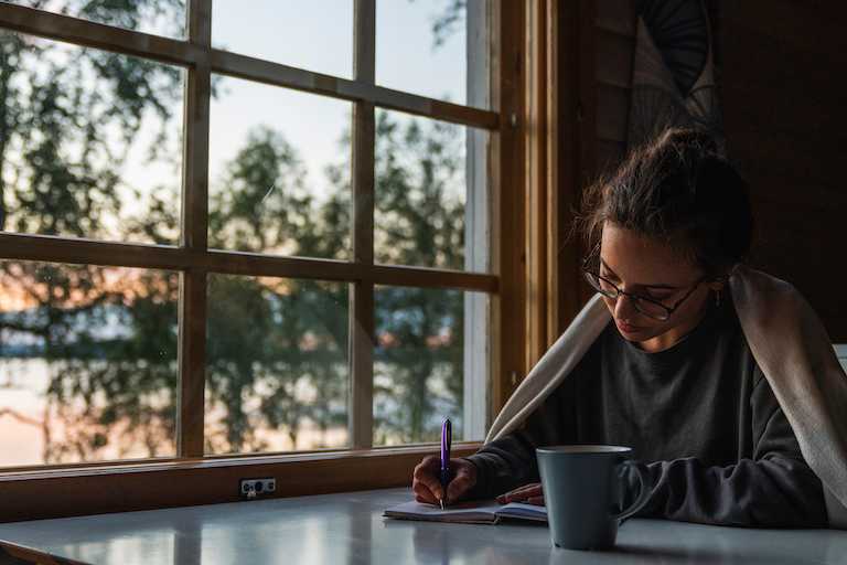 woman with pen, paper, and cup of coffee sits next to window in early morning
