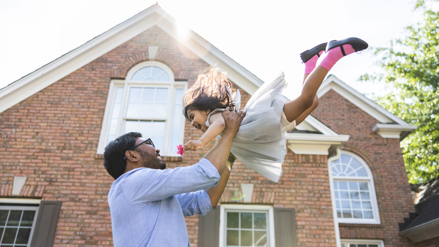 Father lifting toddler daughter in the air in front of suburban home