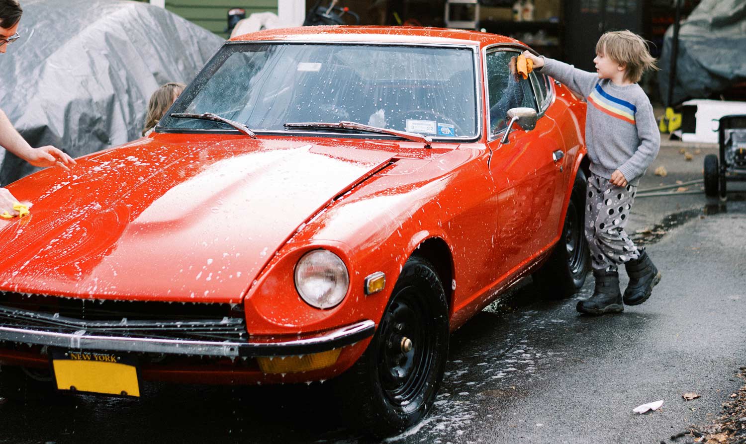 A little boy with his father wash their red vintage car in the driveway of their home