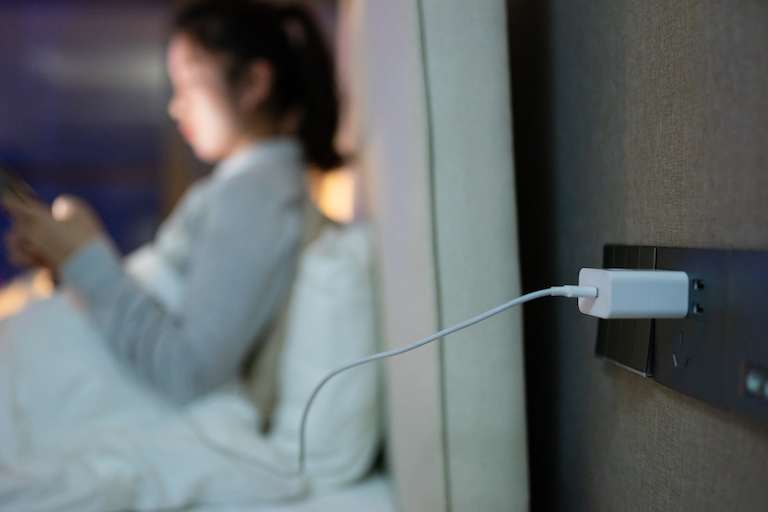 closeup of charger in electrical outlet with woman in bed on her phone blurry in the background