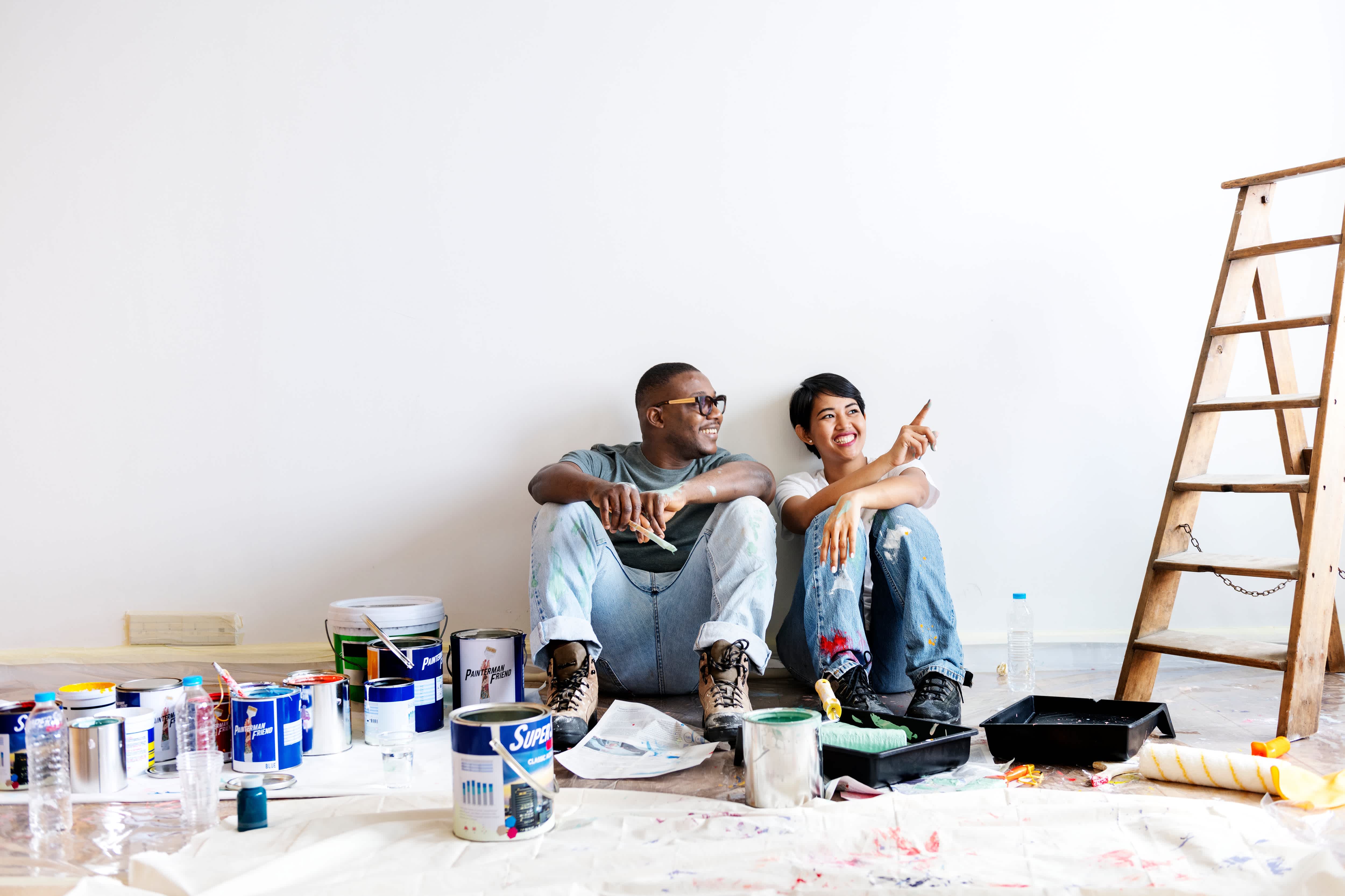 Couple sitting on floor surrounded by paint drop cloths, paint buckets, and a ladder