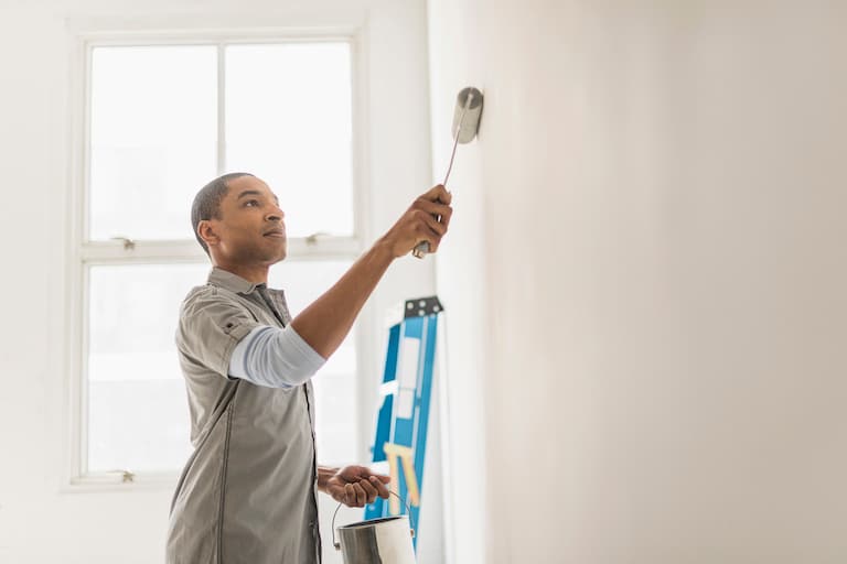 side shot of man painting white wall with white paint, holding roller brush and paint can with blue ladder in background