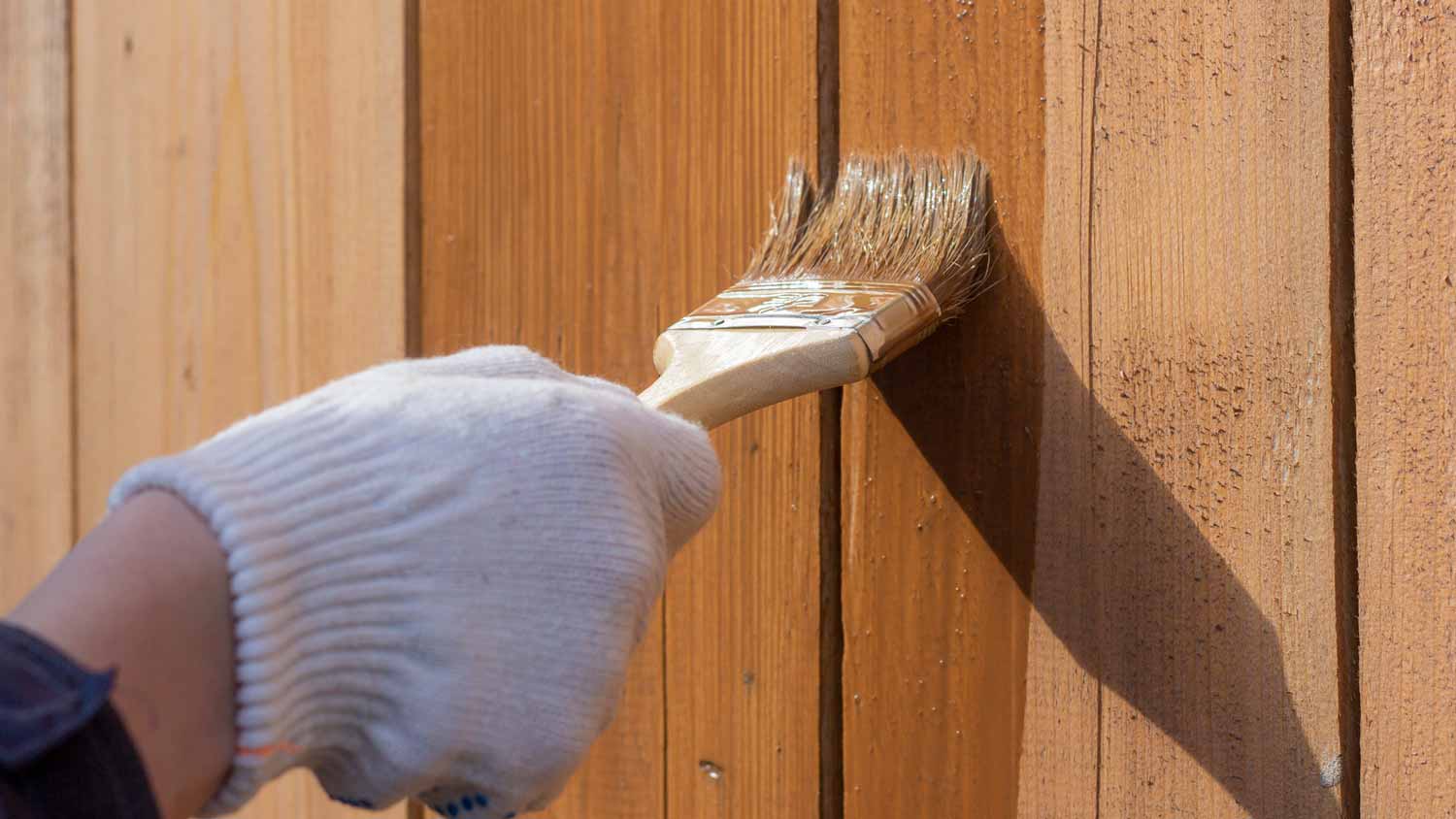 Close-up of a person’s hand in glove sealing a cedar fence