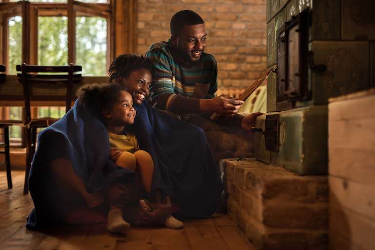 woman, man, and child in cozy blankets huddled next to furnace in low light and smiling