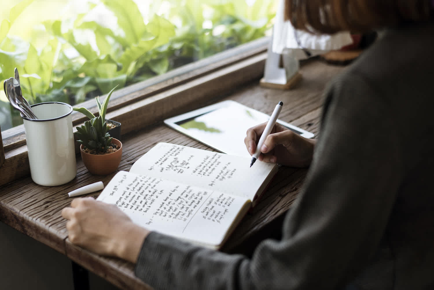 View of young woman with short hair in a blazer writing in her journal on a rustic wooden desk