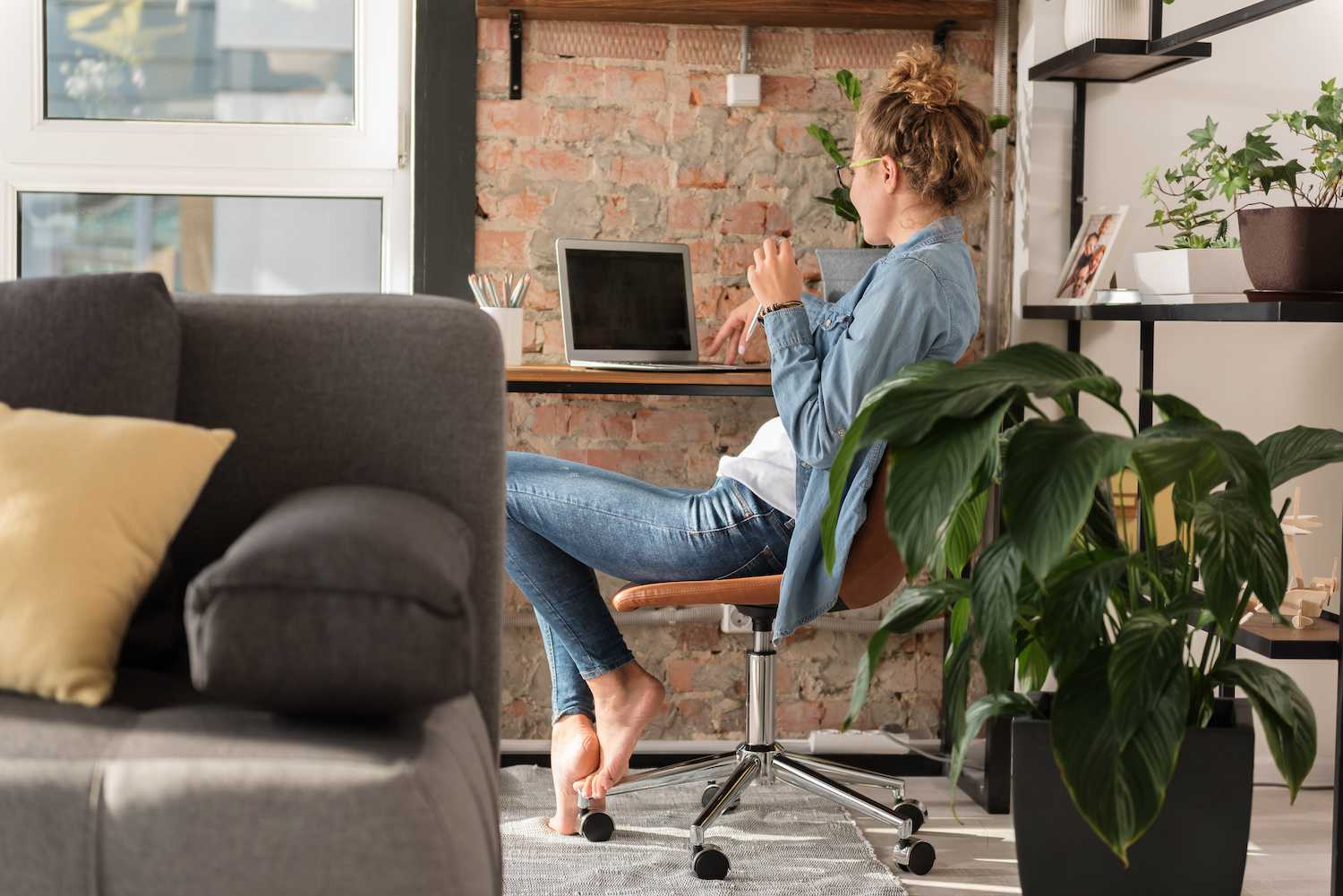 Woman working from home at her desk on her laptop with a large green potted plant next to her desk and plants on the shelves next to her workspace