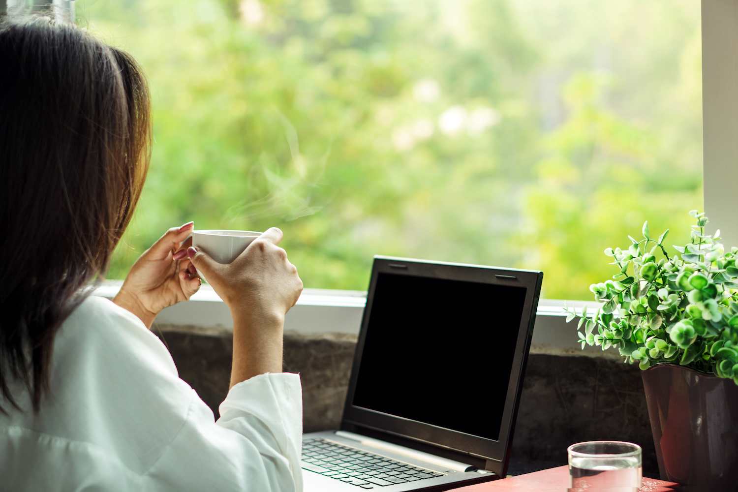 Businesswoman with long black hair drinking coffee at her home office desk facing a window with a view