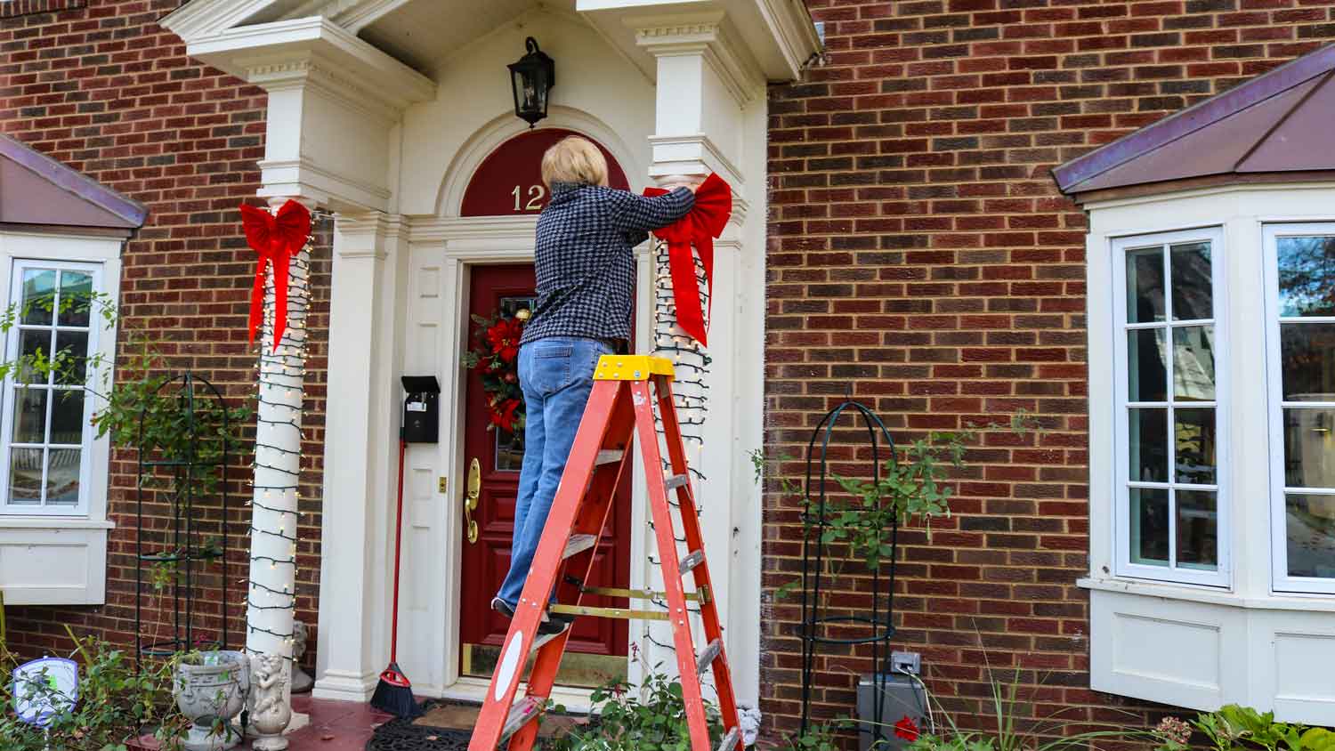 A woman on a ladder hanging Christmas lights outside a brick house