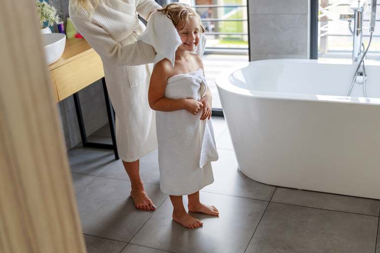 child in white towel gets hair dried by adult in bathroom with gray tiled floors and white bathtub