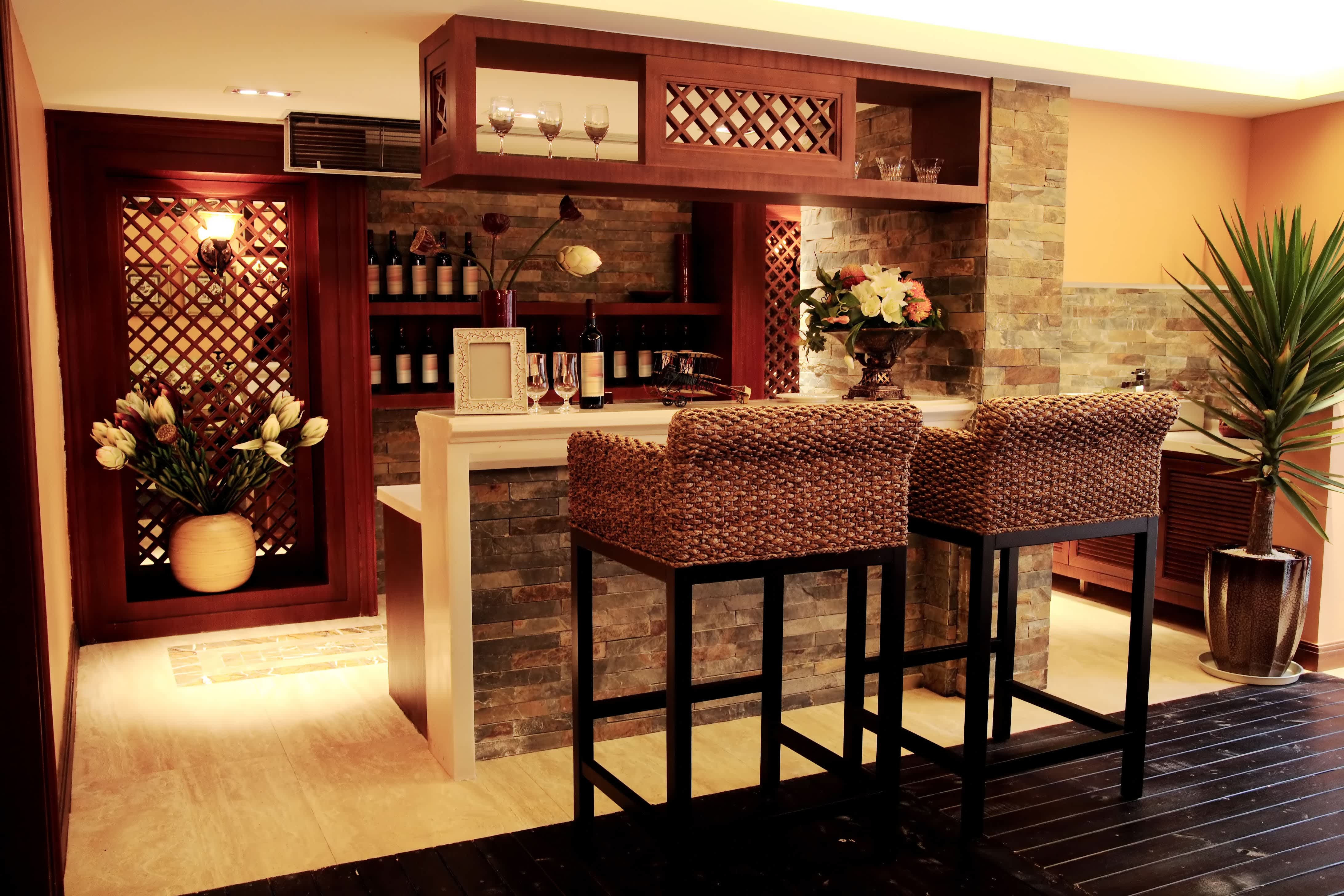 A stylish wet bar in dark wood with wicker seating, brick walls, wine bottles, and plants
