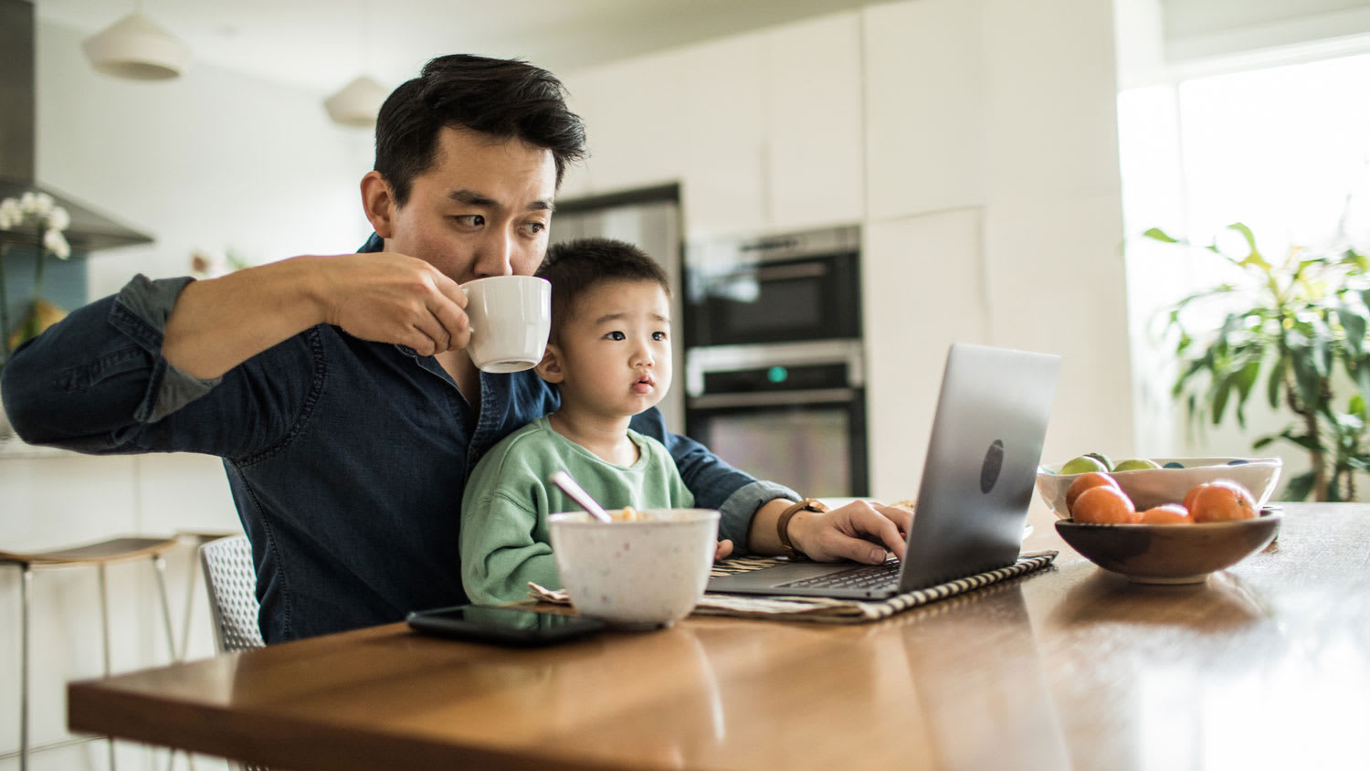 A father multi-tasking with young son on the kitchen table
