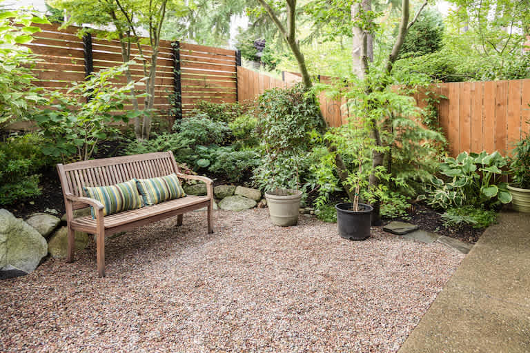 a light wood privacy fence surrounds a private yard with plants, bushes, a bench, and gravel