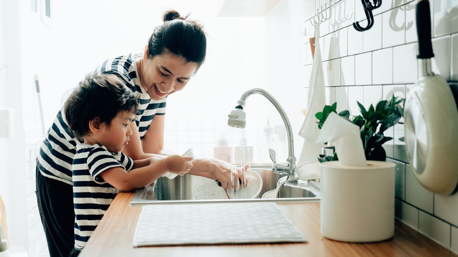 Toddler son helping mom to wash dishes at home