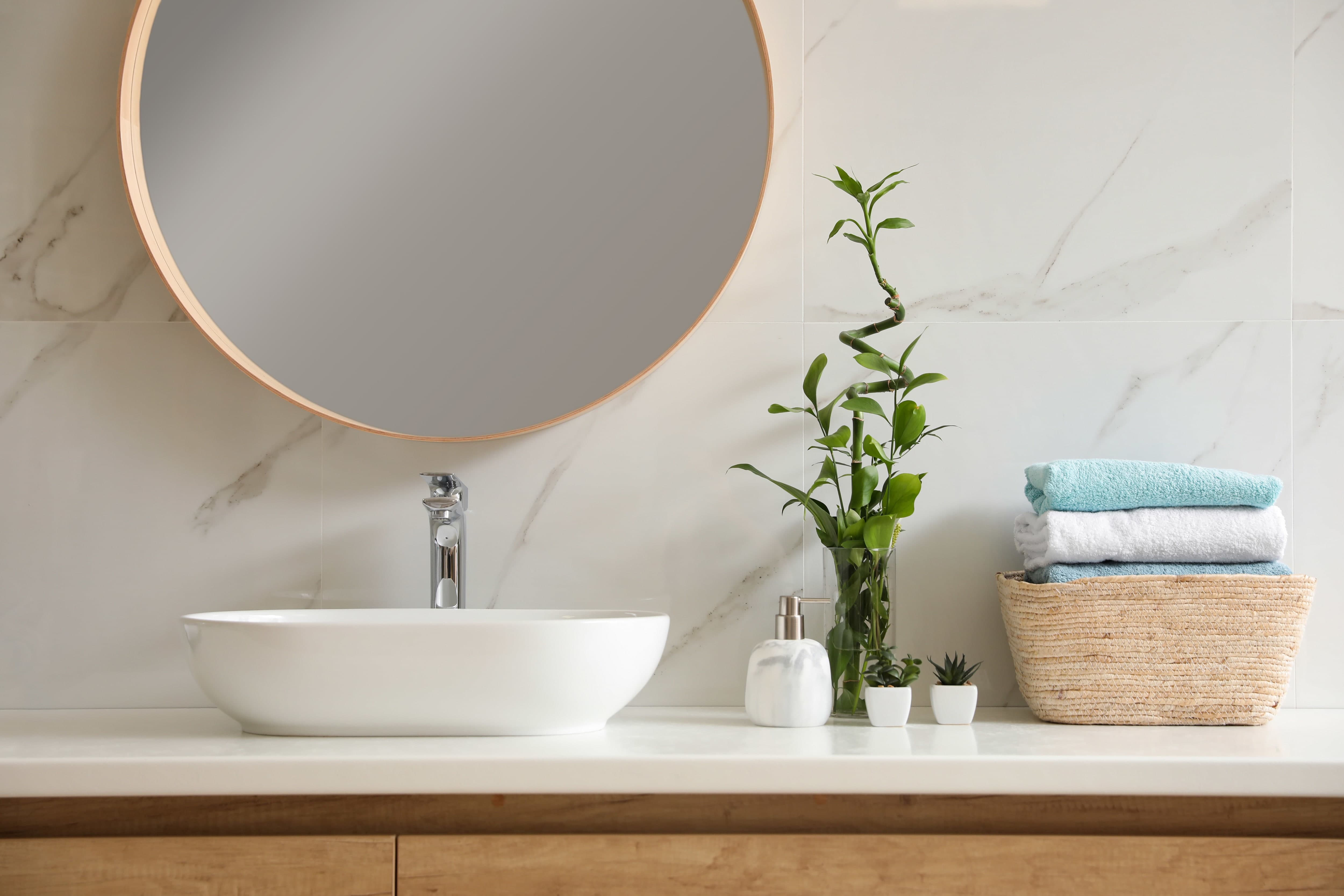 Modern bathroom with marble slab backsplash, round mirror, plants, vessel sink, and a woven basket with towels