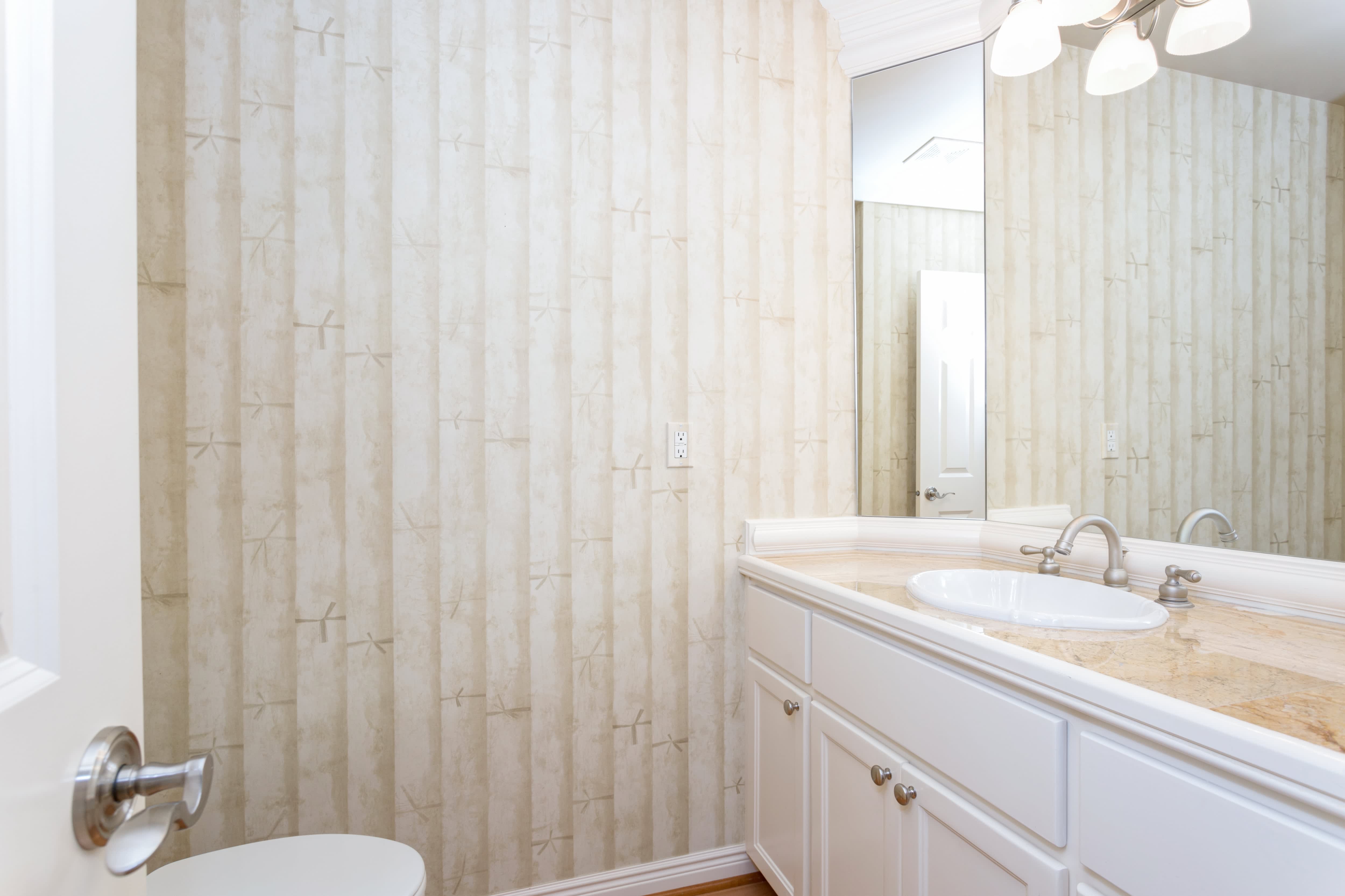 Small white bathroom with marble vanity, a light fixture, a large mirror, and wallpaper featuring wooden designs