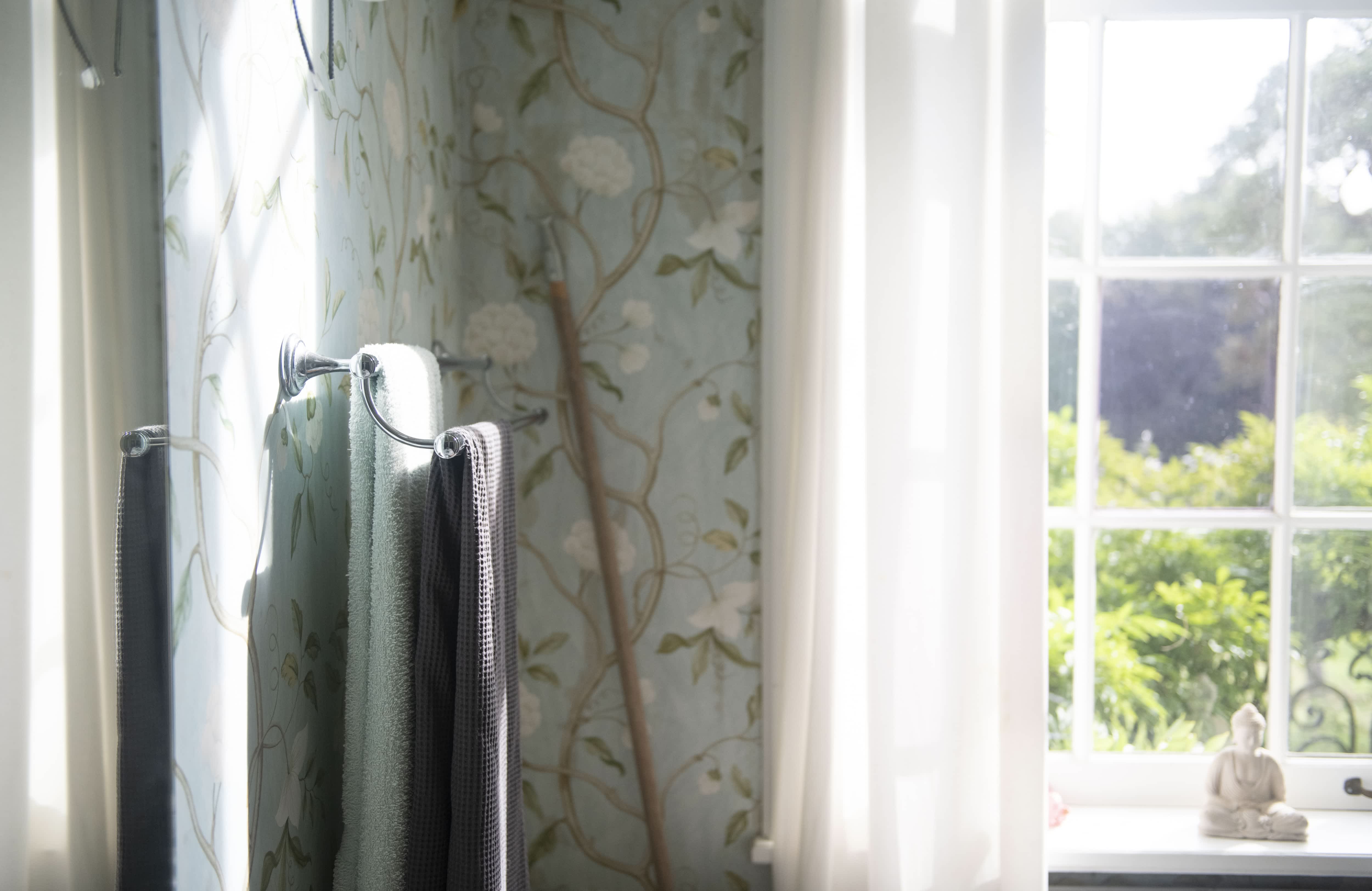 Bright bathroom with floral wallpaper, two towels on a towel rack, a large window, and white curtains
