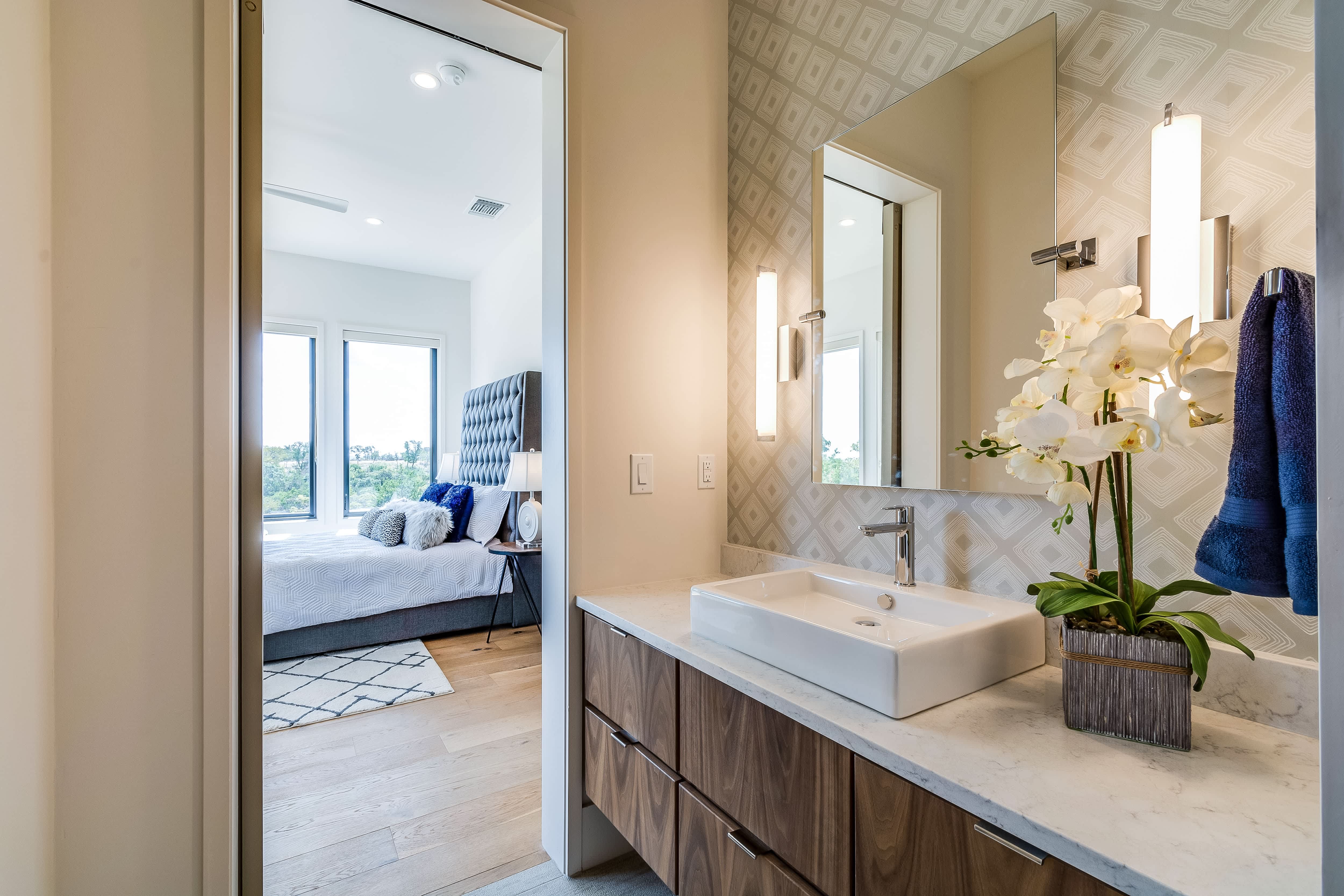 En suite bathroom with wooden vanity, mirror, vessel sink, and geometric wallpaper overlooking a modern bedroom