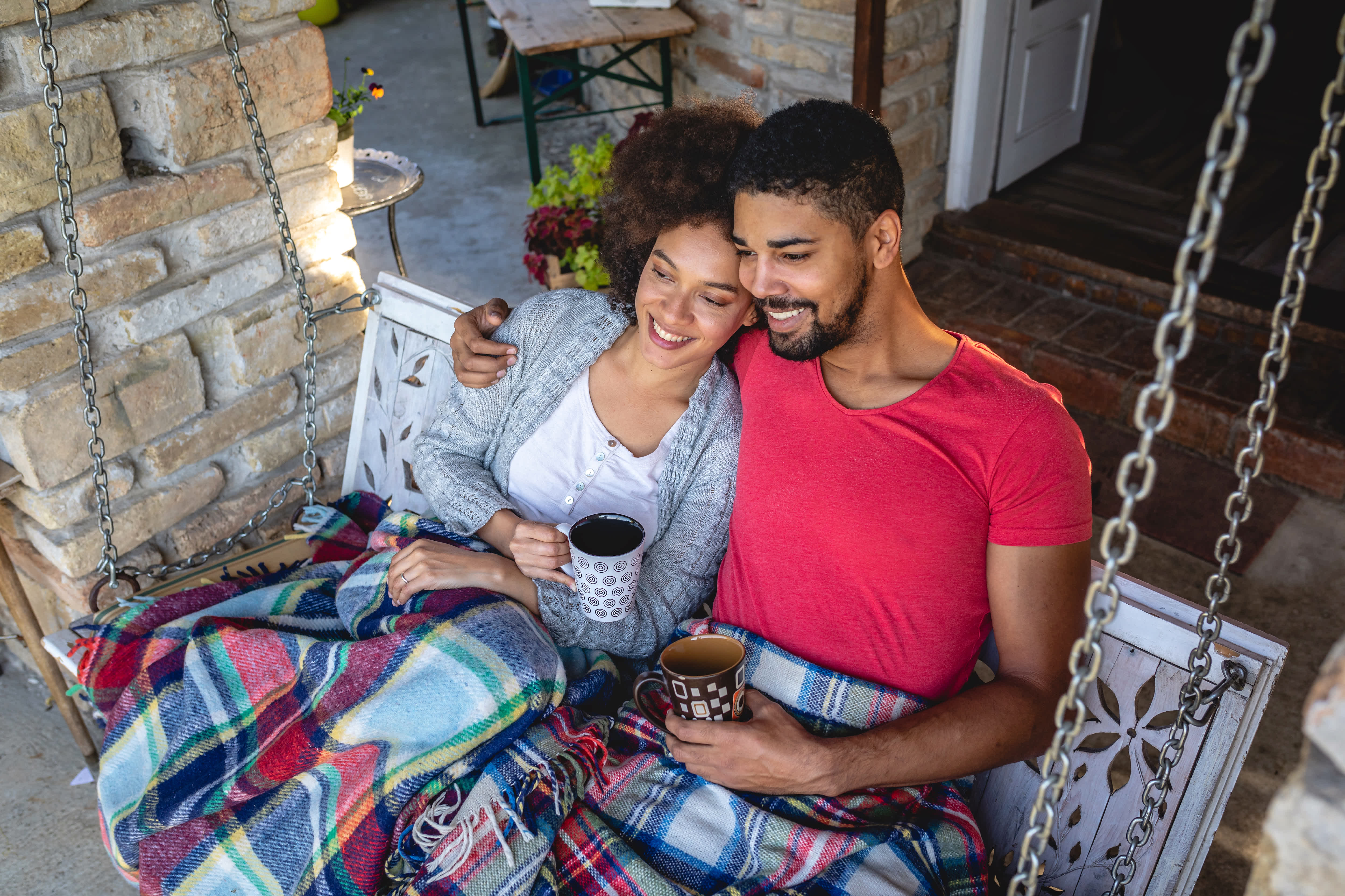 A couple on a porch swing under blankets holding mugs