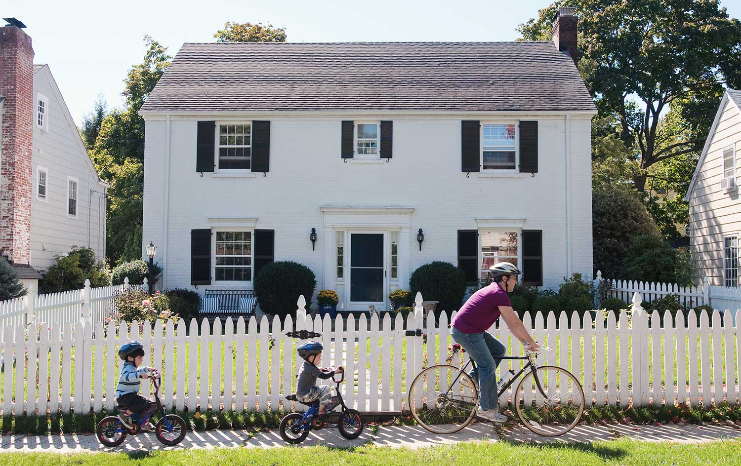 Father and kids riding bikes in front of fenced house